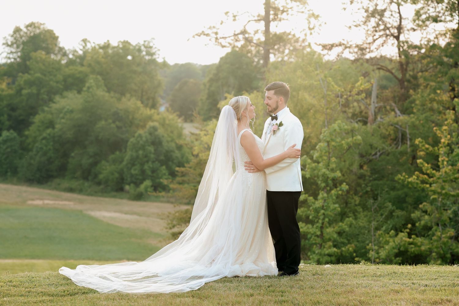Bride and groom share romantic sunset portrait on green hillside, cathedral veil trailing behind.