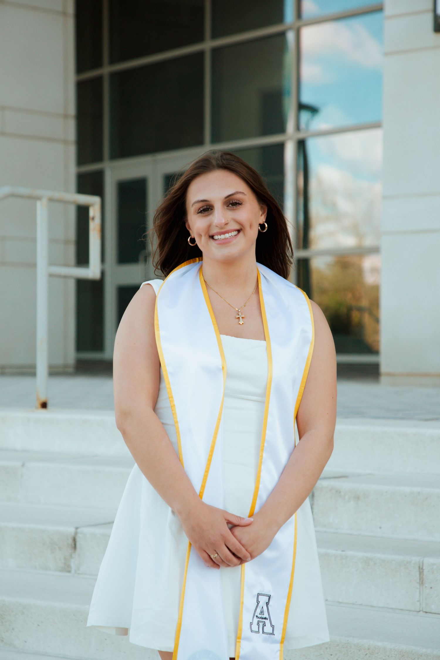 Smiling graduate in white dress and gold stole poses on steps outside university building.