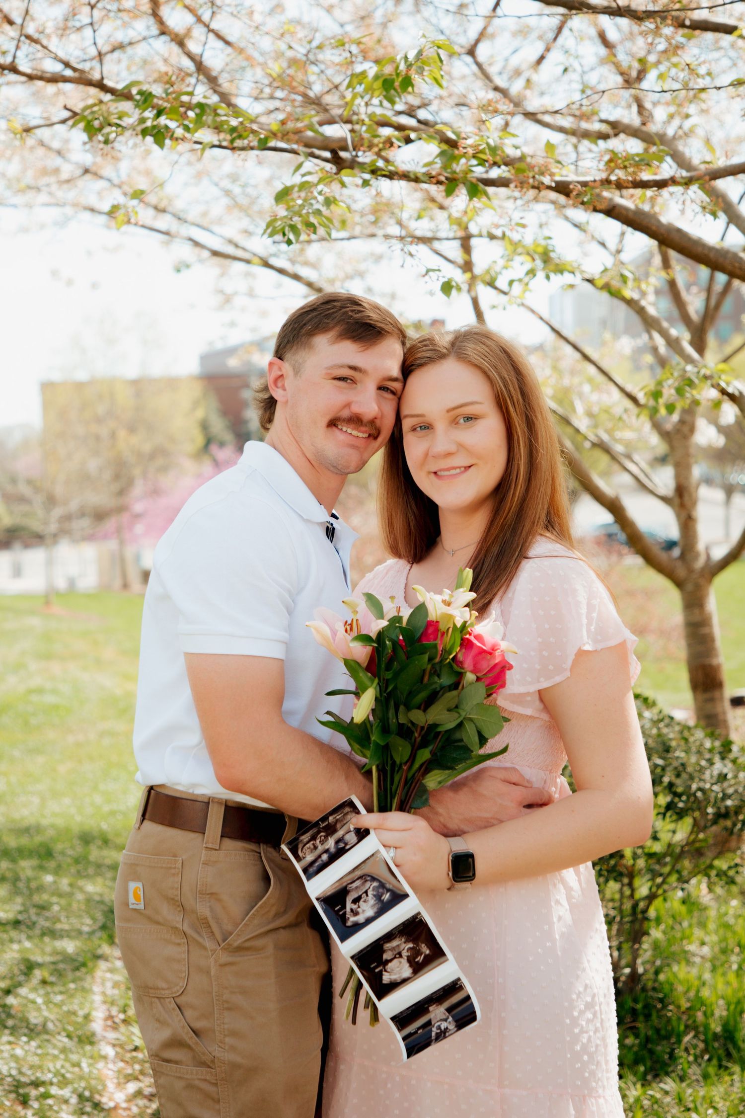 Happy expecting couple posing under cherry blossom tree holding flowers and ultrasound strip.