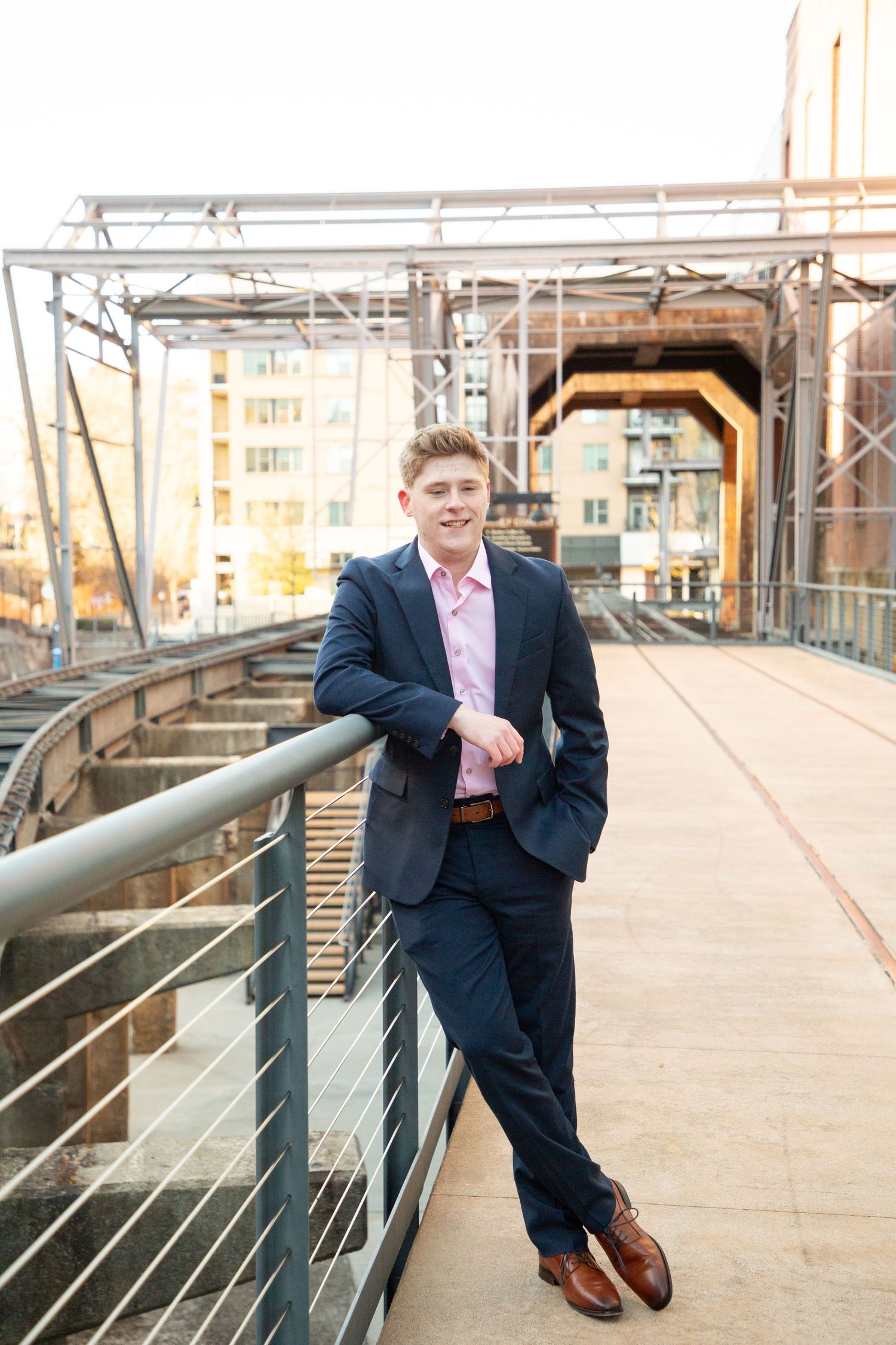 Smiling young man in navy suit and pink shirt posing on an industrial bridge railing.