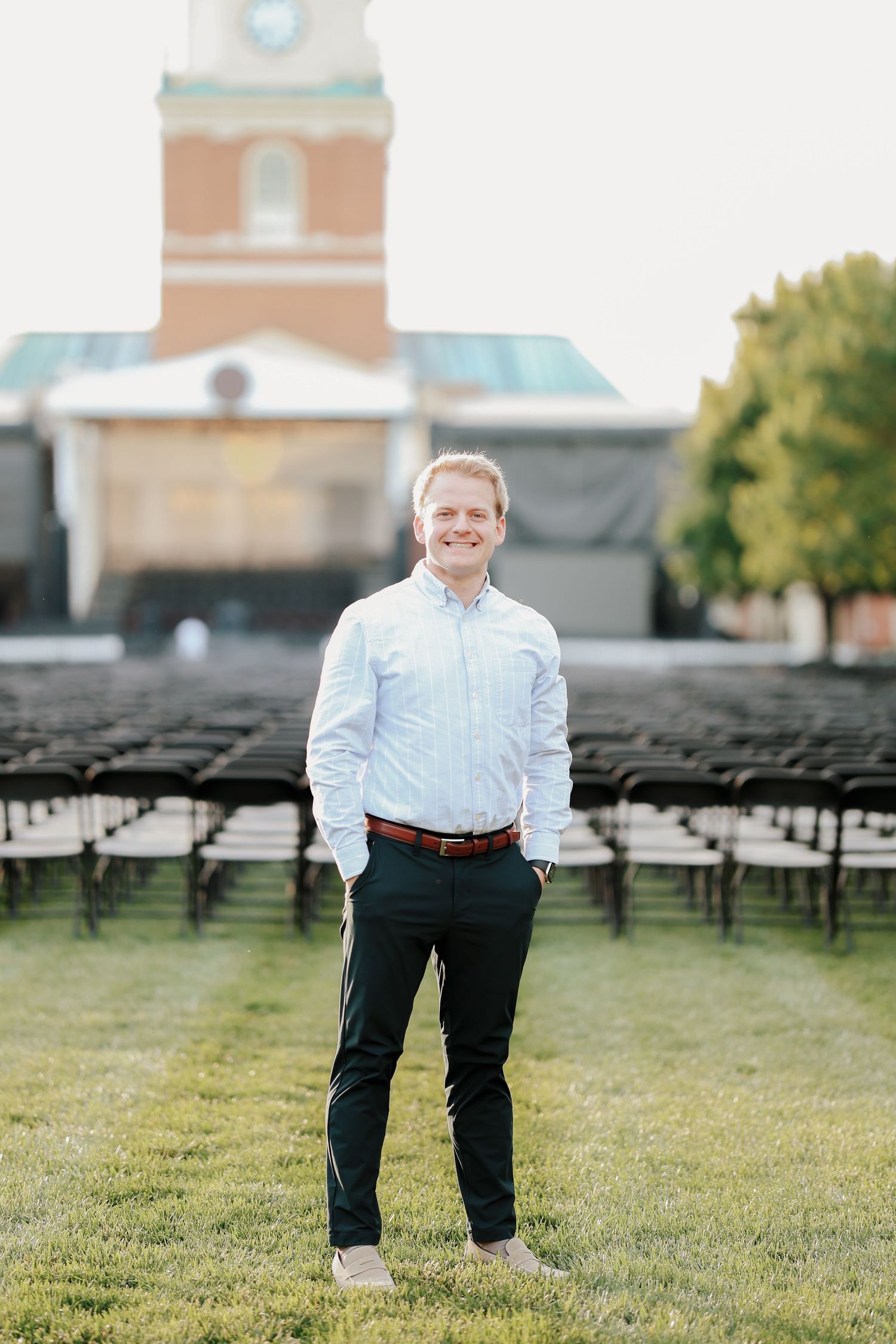 Young man in light blue shirt and dark pants smiling outdoors near rows of chairs and brick building.