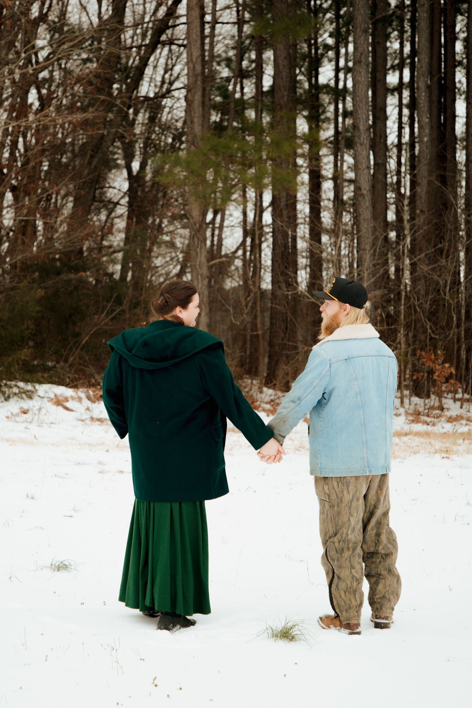 Couple walking hand in hand through snowy path toward winter forest, seen from behind.