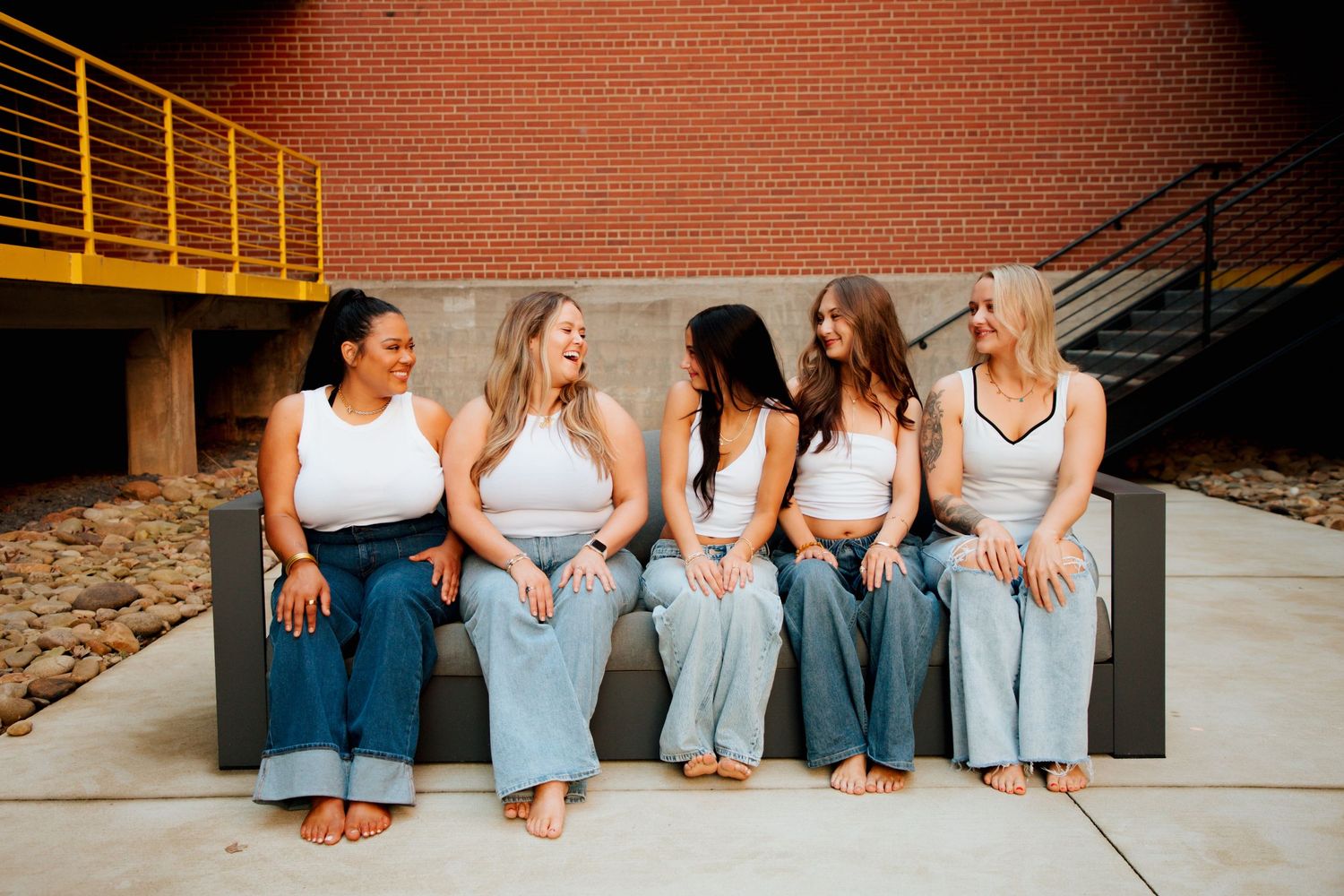 Five women in white crop tops and blue jeans sitting together on a bench outdoors near a brick wall.
