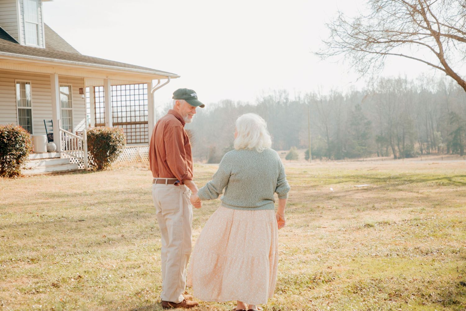 Elderly couple holding hands while walking across a sunlit lawn beside a classic white farmhouse.