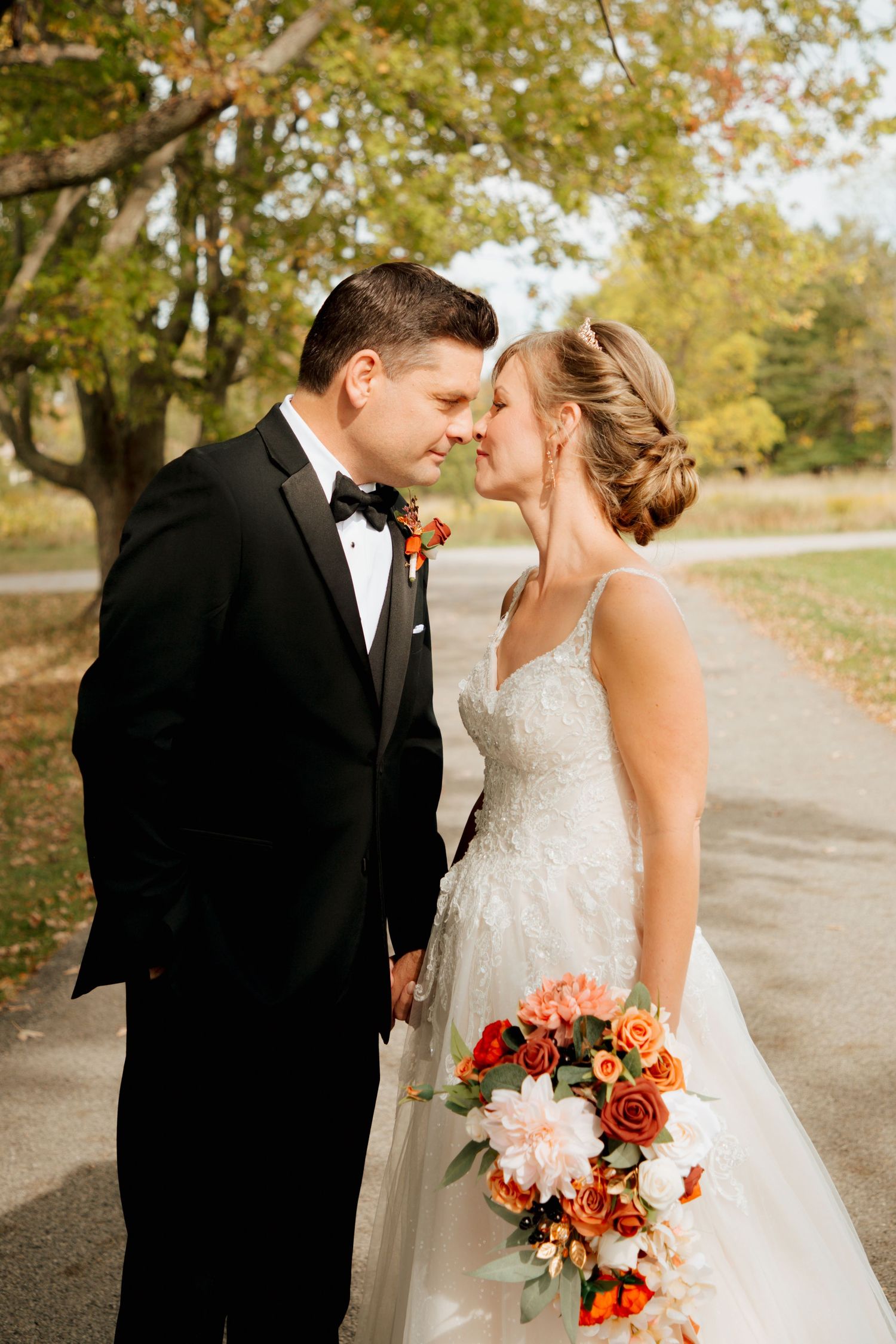 Bride and groom touching foreheads tenderly, holding fall floral bouquet in autumn setting.