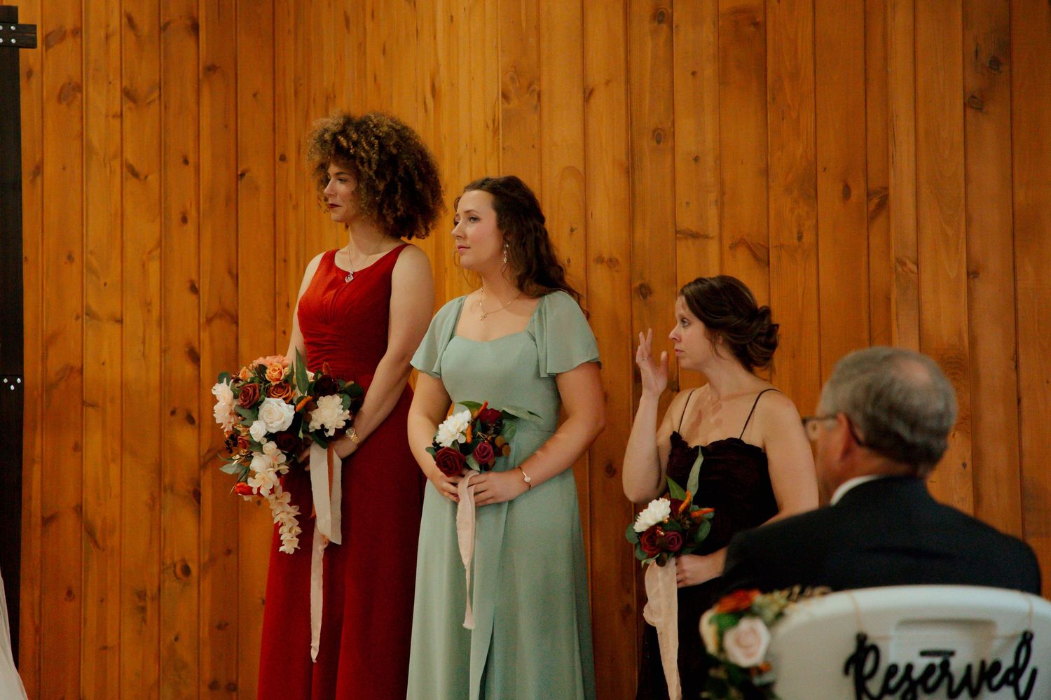 Three bridesmaids in red, sage green, and black dresses holding bouquets at wedding ceremony.
