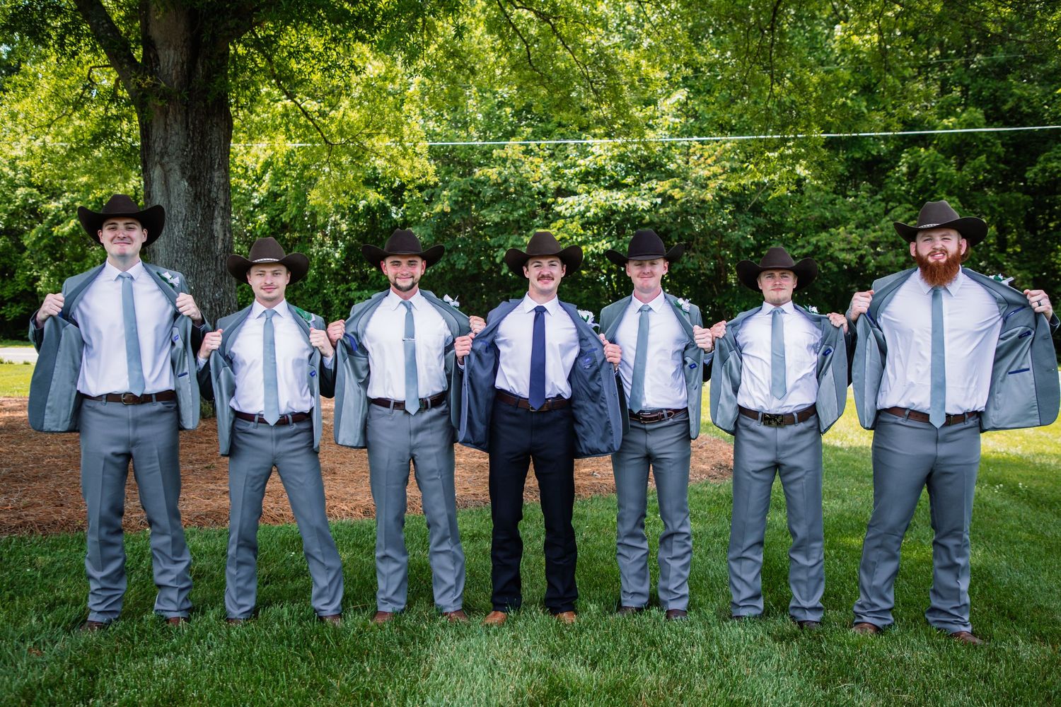 Groomsmen in gray suits and cowboy hats open their jackets in a fun pose at an outdoor country wedding.