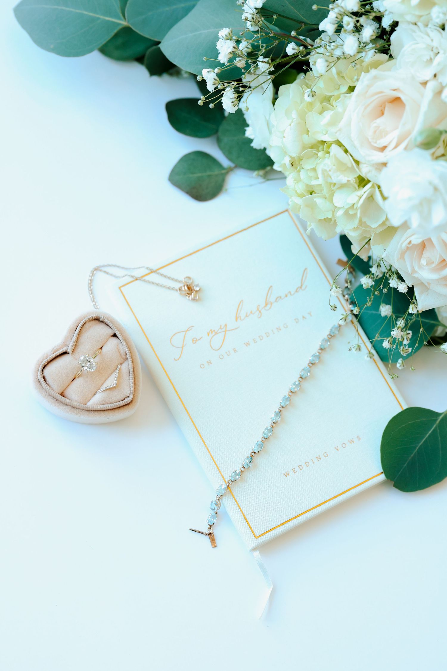 Wedding flat lay with bridal bouquet, ring box, necklace, and vow book on white background.