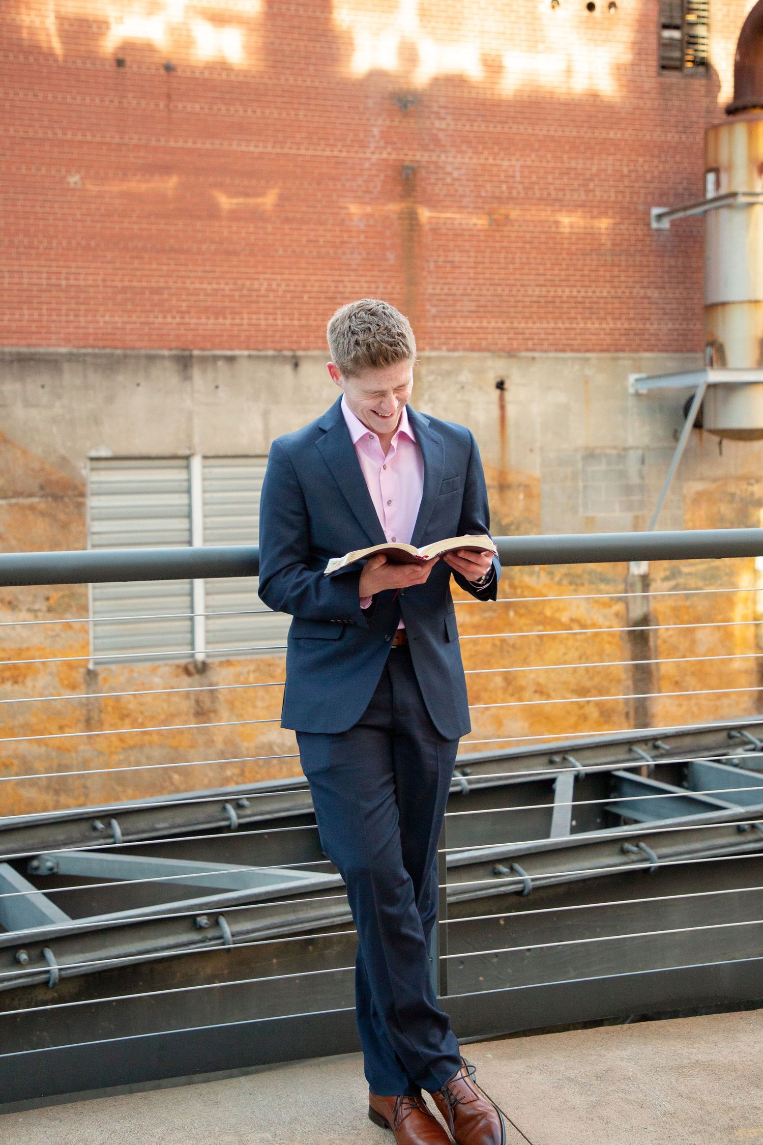 Young man in navy suit reading a book while leaning on a metal railing outdoors.