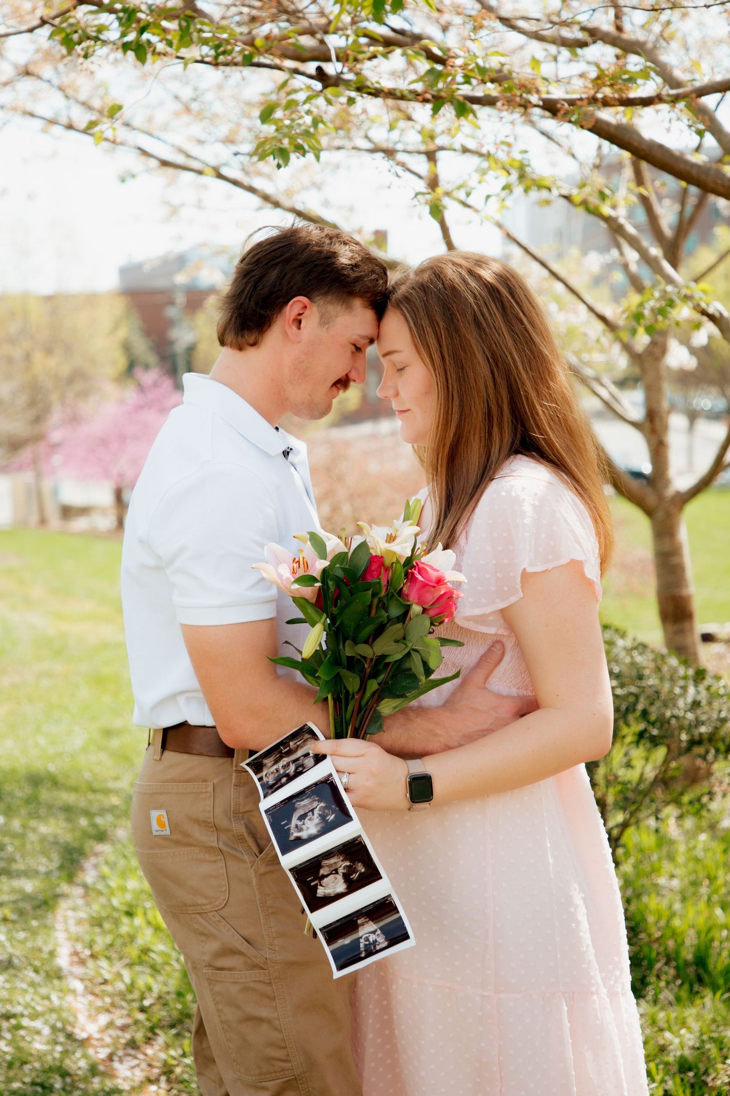Expecting couple embracing under blooming tree holding flowers and ultrasound photos.