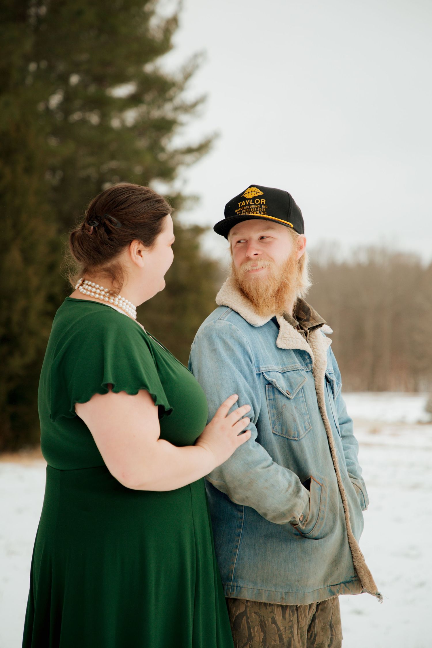Bearded man in denim jacket and cap with woman in green dress sharing a tender moment in snow.