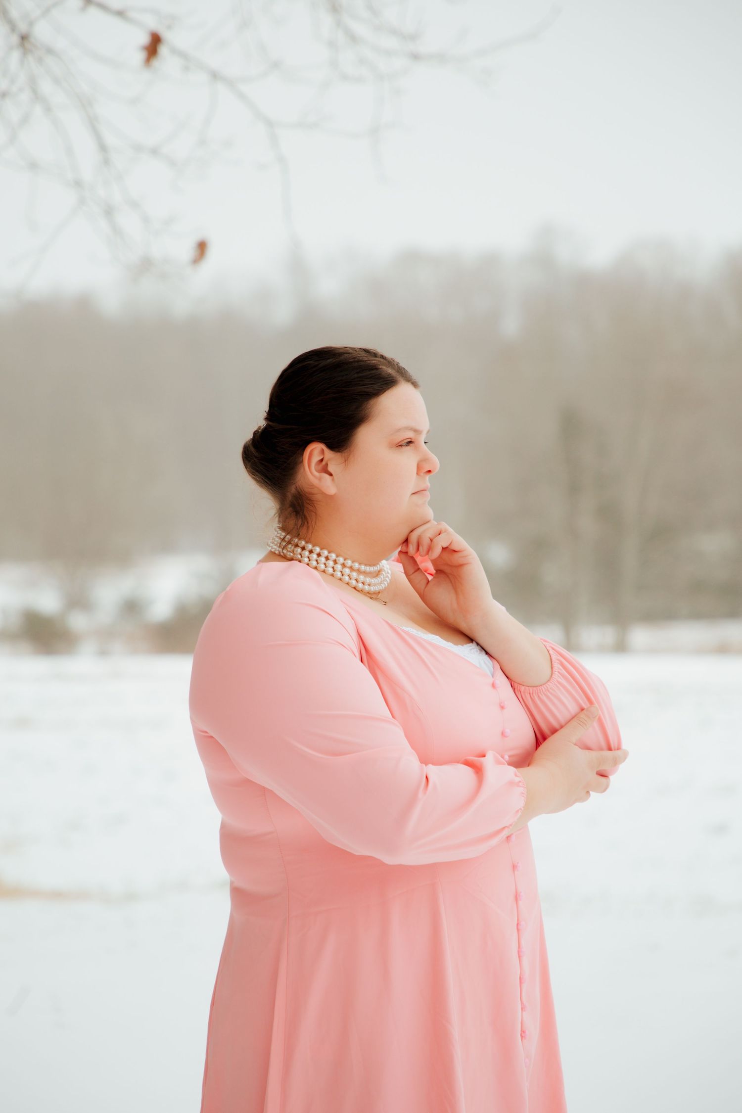 Plus-size woman in pink dress with pearl necklace posing thoughtfully in a snowy field.