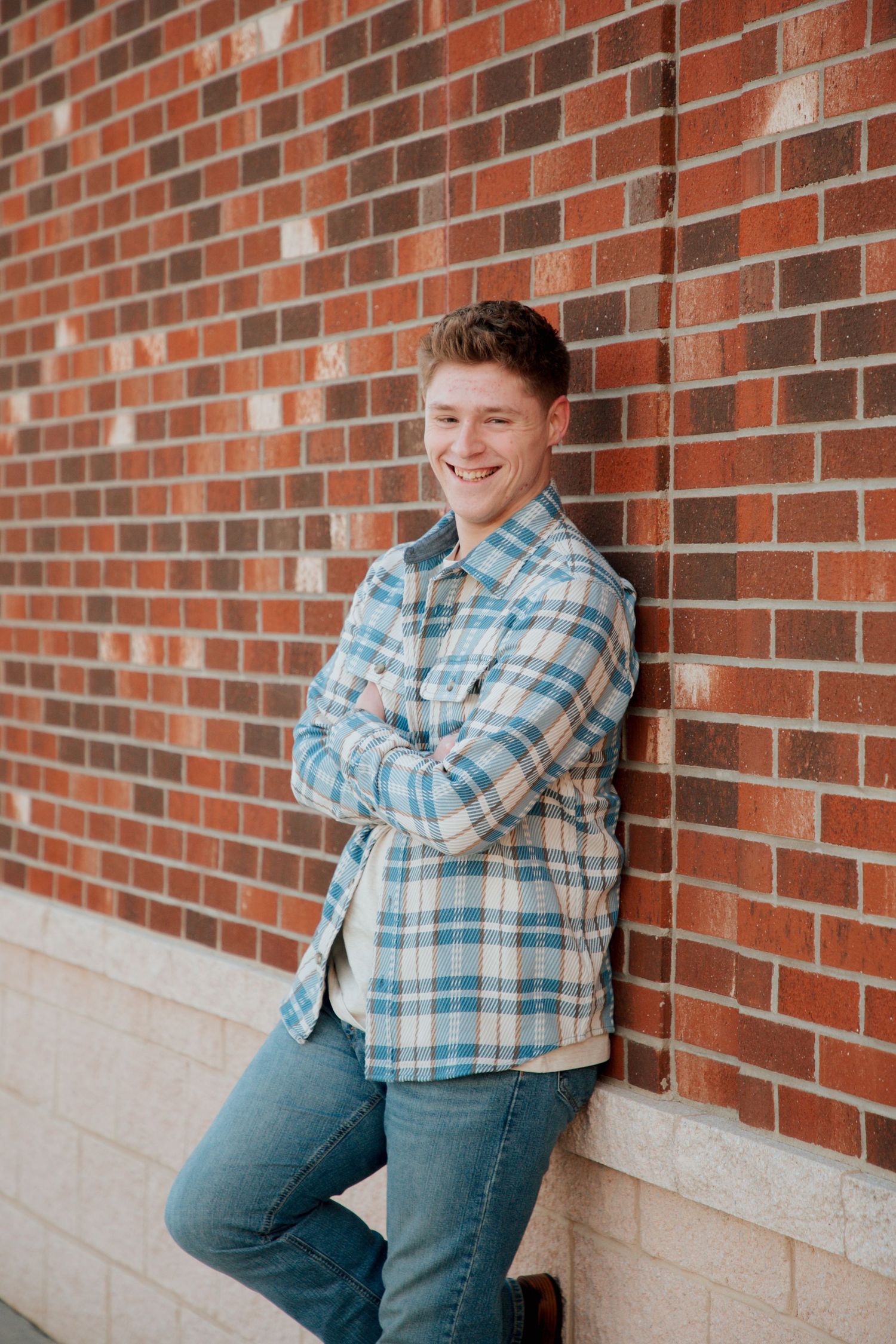 Young man in blue plaid shirt smiling with arms crossed against red brick wall.