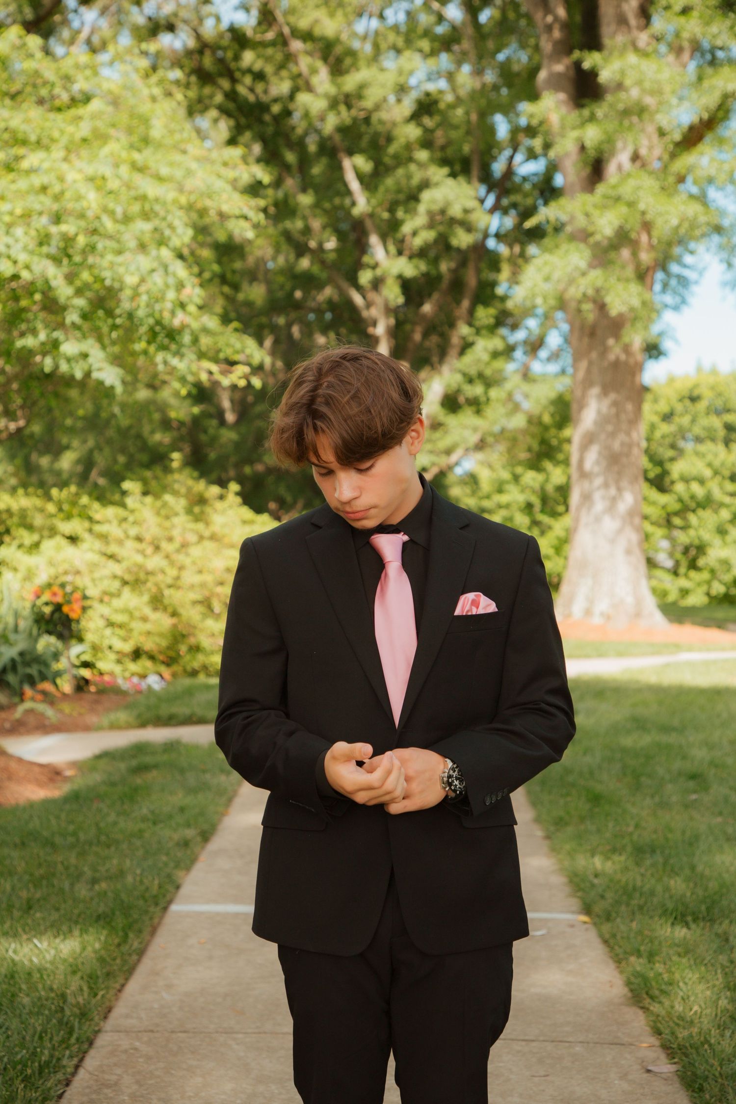 Young teen boy in black suit with pink tie and pocket square stands on garden pathway.