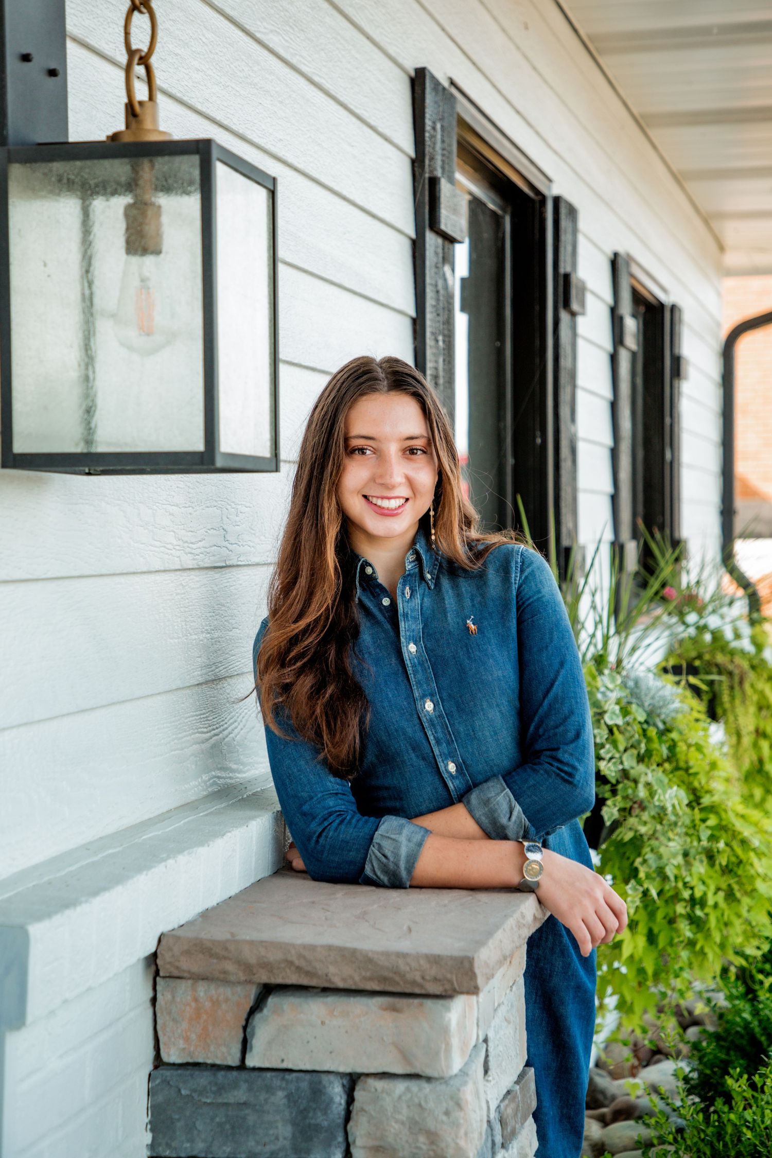 Smiling young woman in denim shirt leaning on stone pillar of white farmhouse porch with plants.