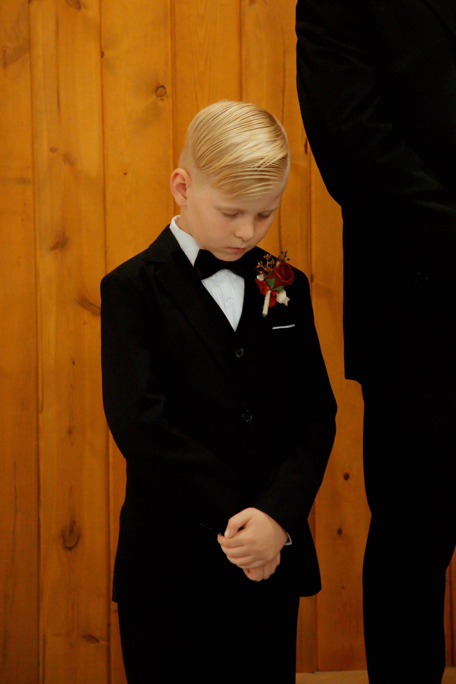 A young blonde ring bearer in a black tuxedo with boutonniere stands with hands clasped during a wedding.