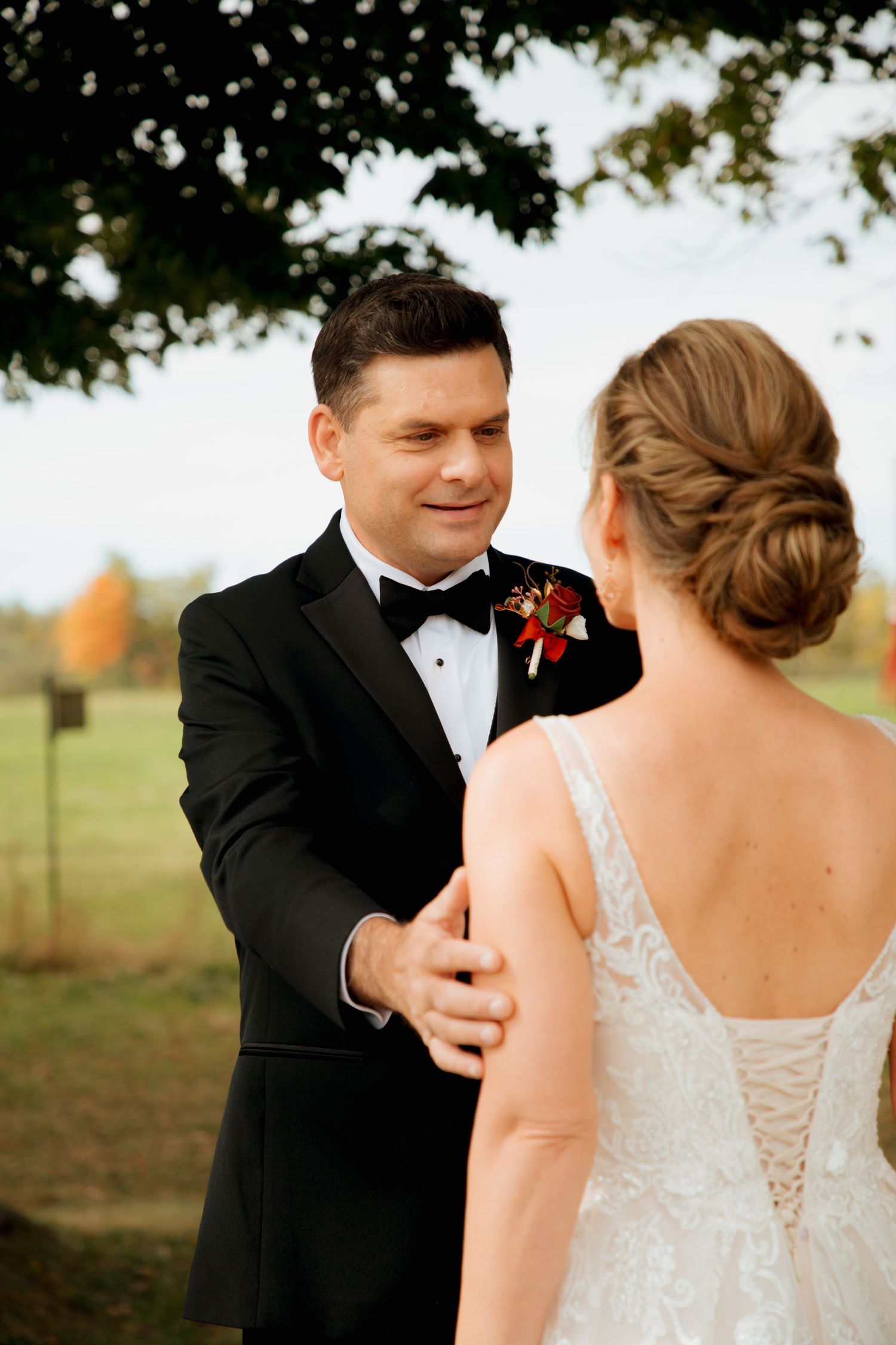Groom in black tuxedo smiling at bride during outdoor fall wedding first look ceremony.