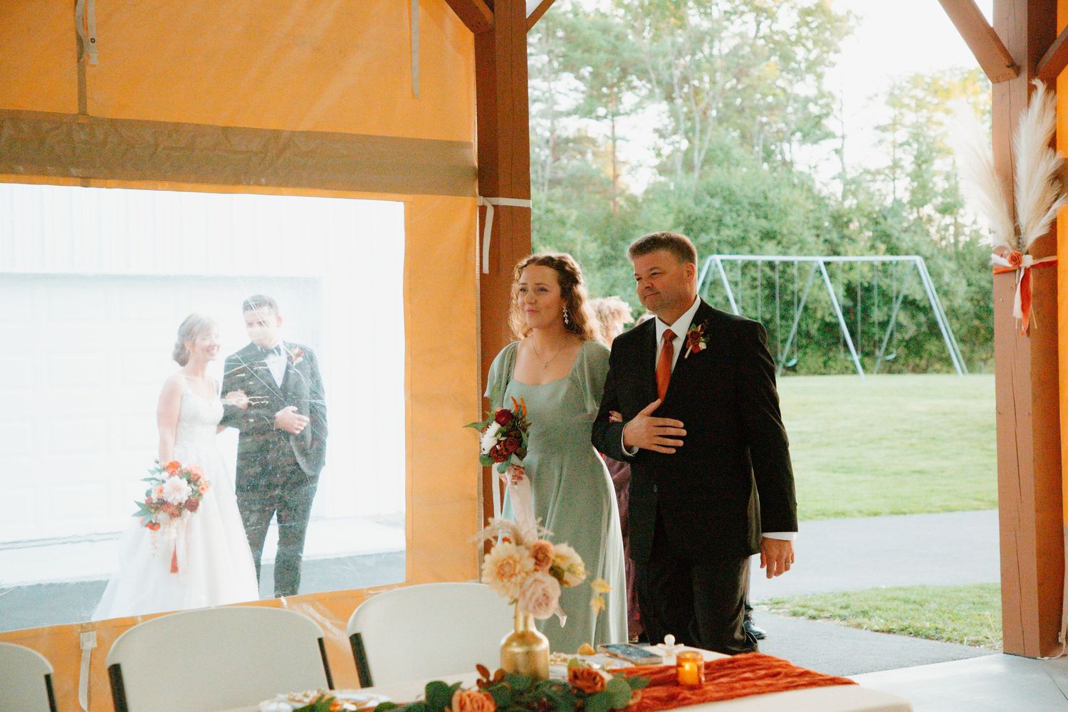 A bridesmaid in sage green escorts a guest during a rustic outdoor wedding ceremony under a wooden pavilion.