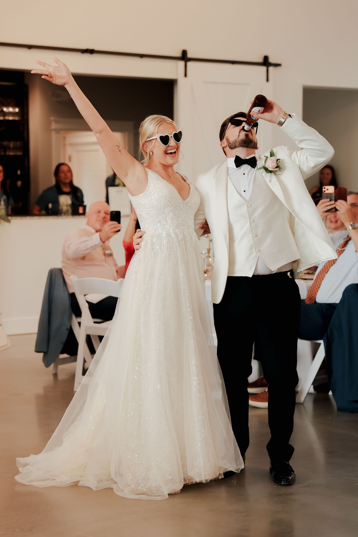 Bride and groom in sunglasses celebrating joyfully during wedding reception grand entrance in bright white barn venue.