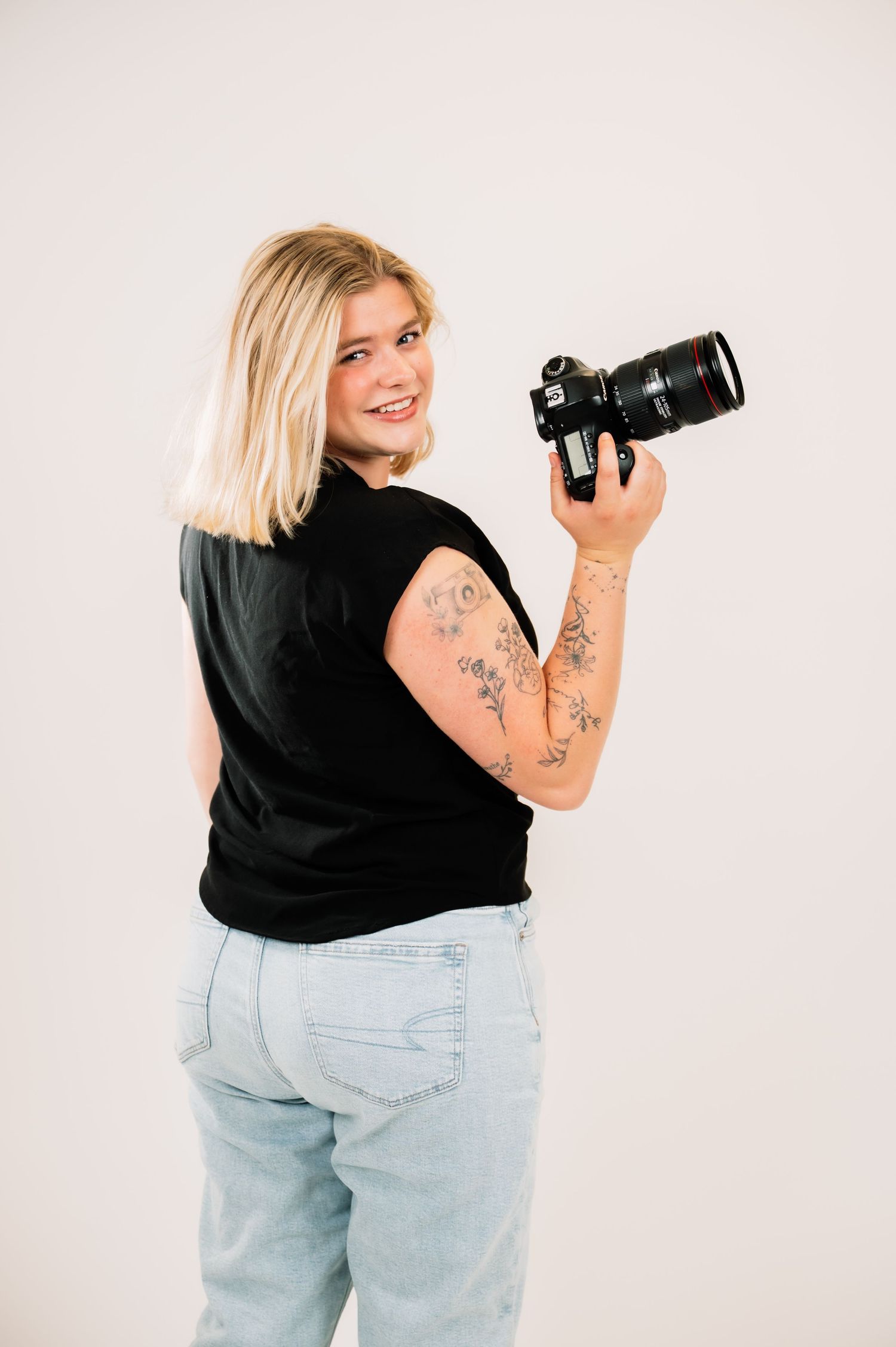 Blonde female photographer turning to smile while holding Canon DSLR camera with floral arm tattoos.