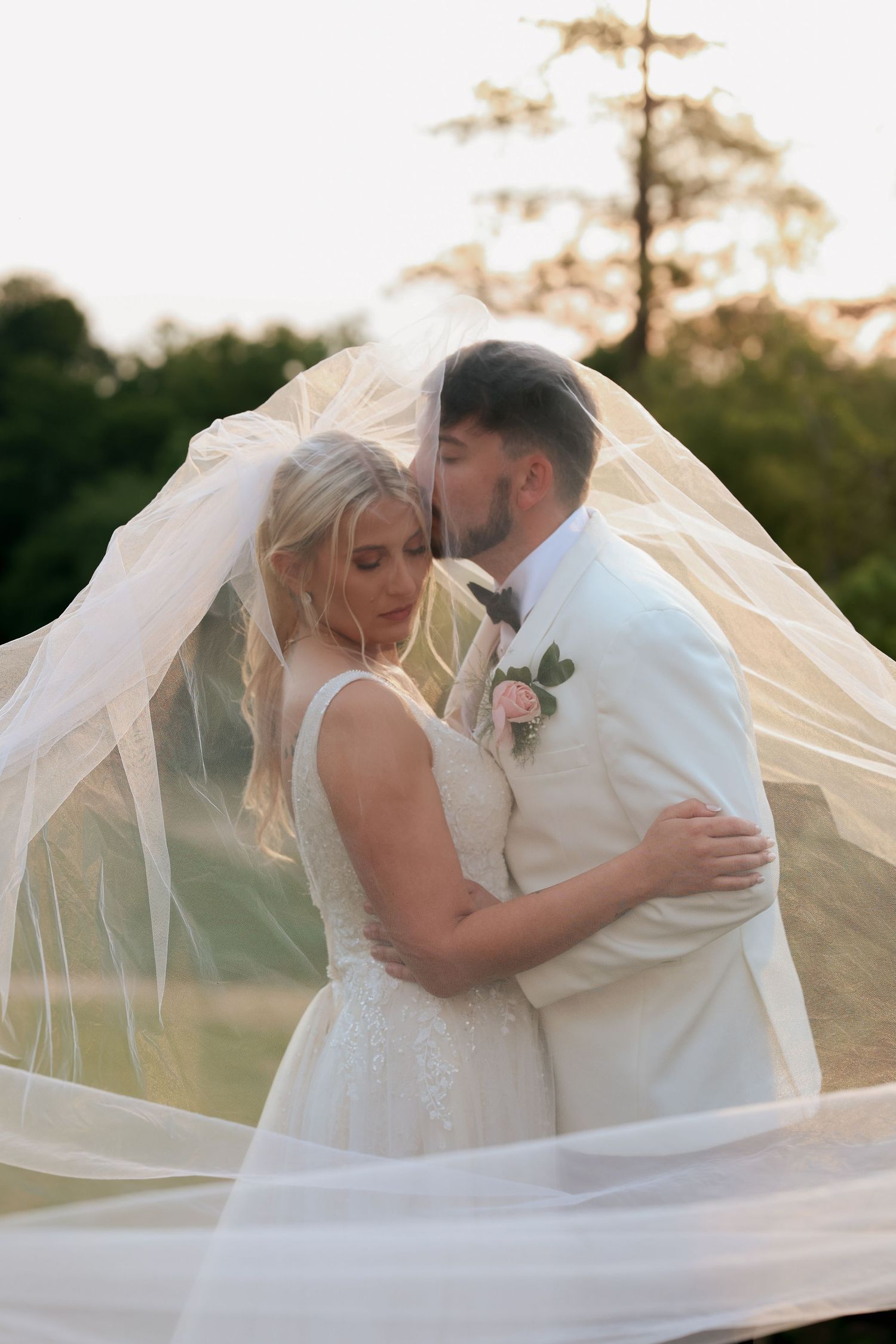 Groom kisses bride on cheek under flowing veil at golden hour during outdoor wedding portrait session.