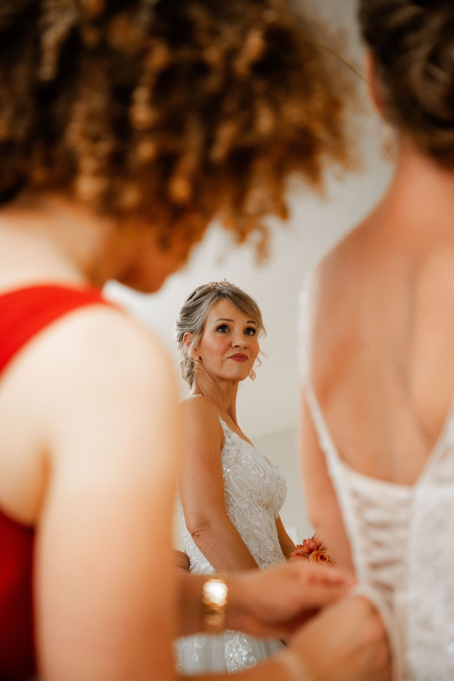 Bride smiling in mirror reflection while bridesmaids help lace her wedding dress.