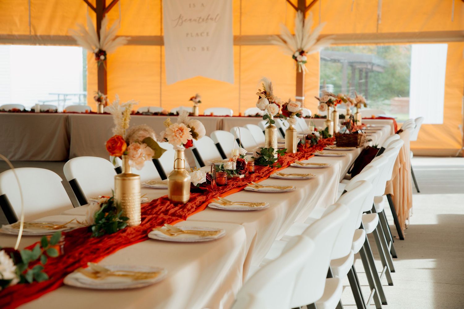 Elegant wedding banquet table with red runner, gold floral centerpieces, white plates, and decorative banner in tent venue.