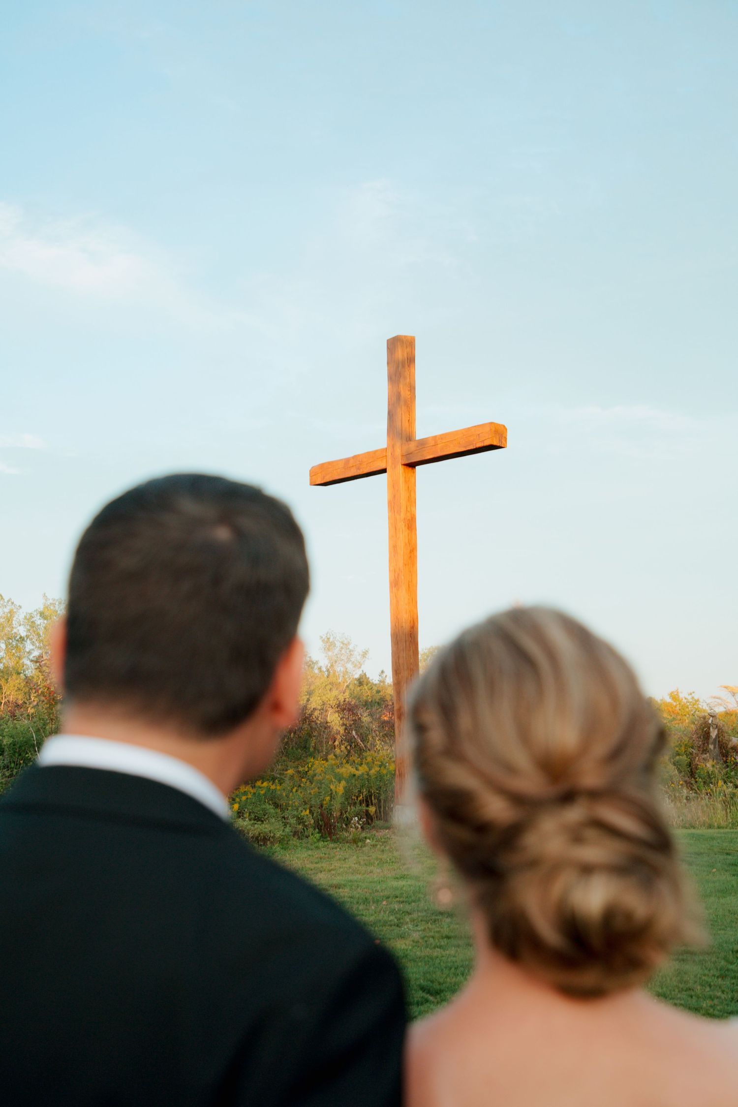Bride and groom viewed from behind looking at a tall wooden cross in an open field.