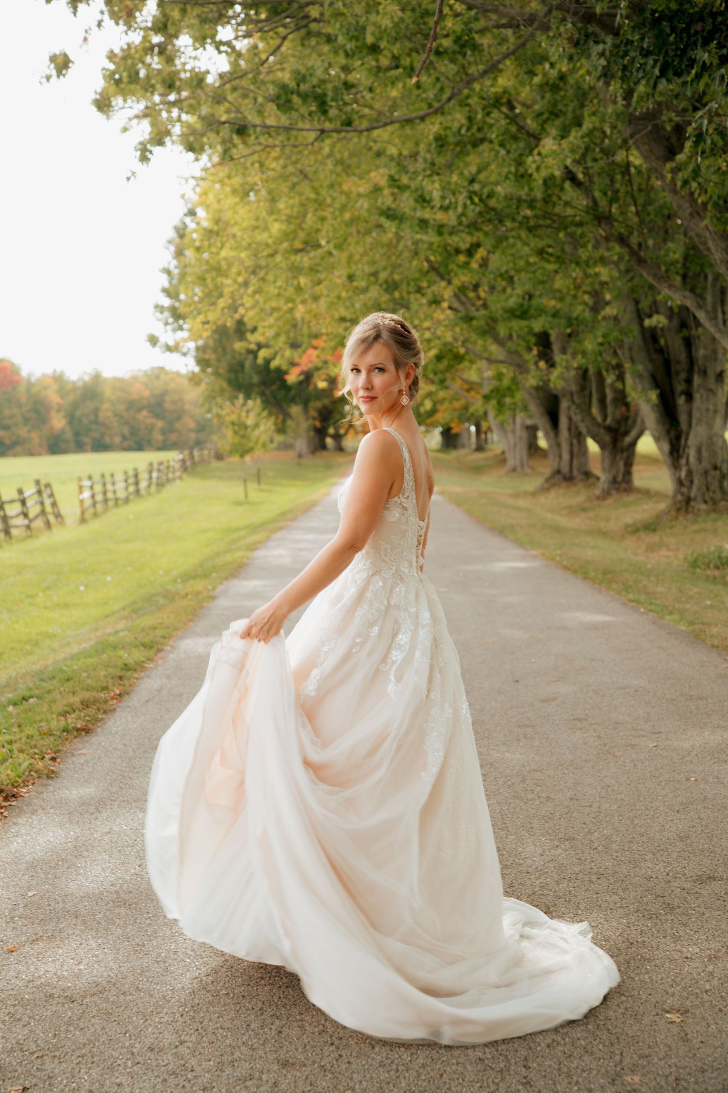 Bride in a flowing white gown twirling on a tree-lined path during an autumn outdoor wedding.