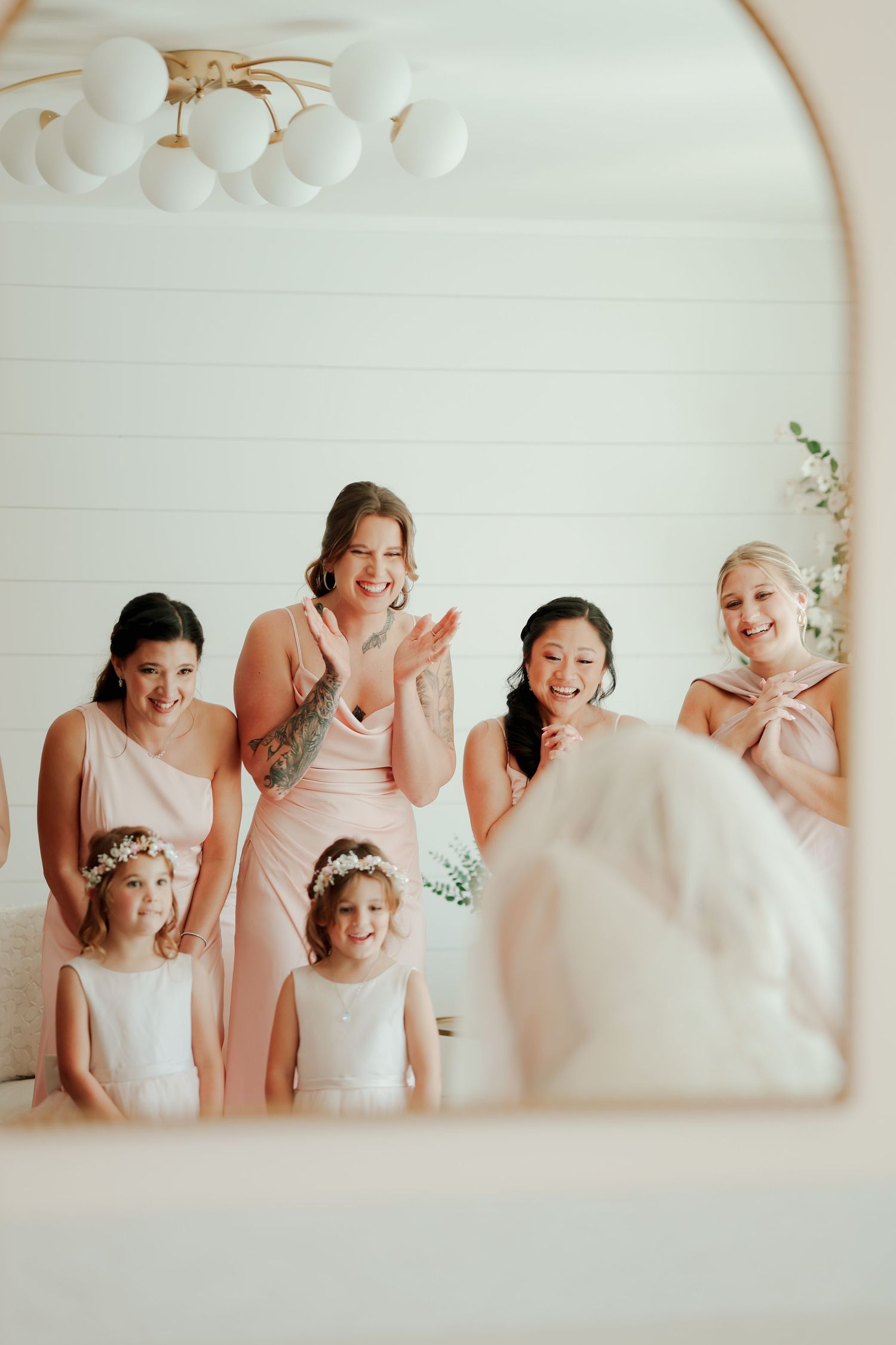 Bridesmaids and flower girls in blush dresses react with joy seeing bride in mirror reflection.