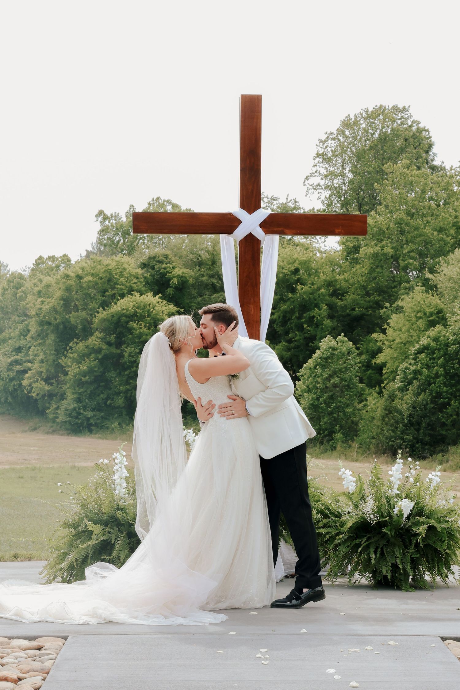 Bride and groom share first kiss in front of a large wooden cross at outdoor ceremony.
