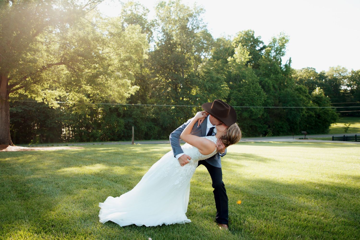 Groom dipping bride in white wedding gown for a romantic kiss on sunlit green lawn.