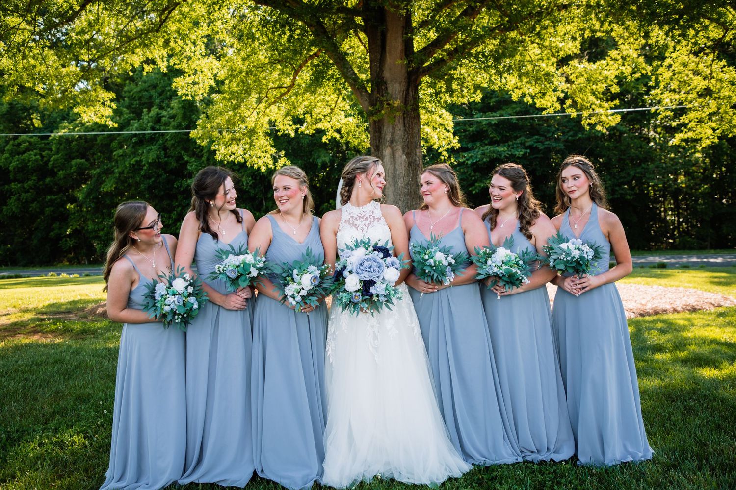 Bride surrounded by bridesmaids in dusty blue gowns holding white and green bouquets outdoors under a large tree.