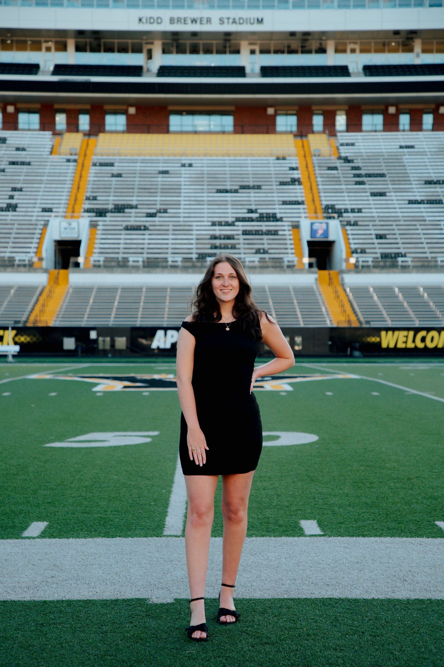 Female graduate in black dress standing on Kidd Brewer Stadium football field at Appalachian State University.