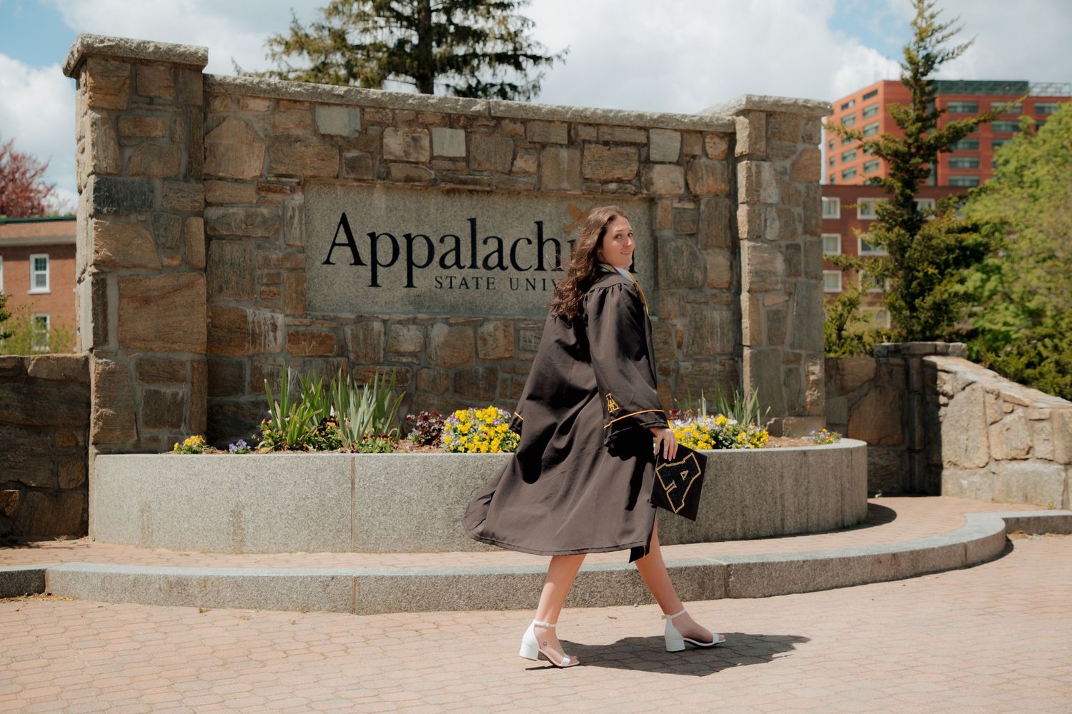 Female graduate in black gown walking past Appalachian State University stone entrance sign.