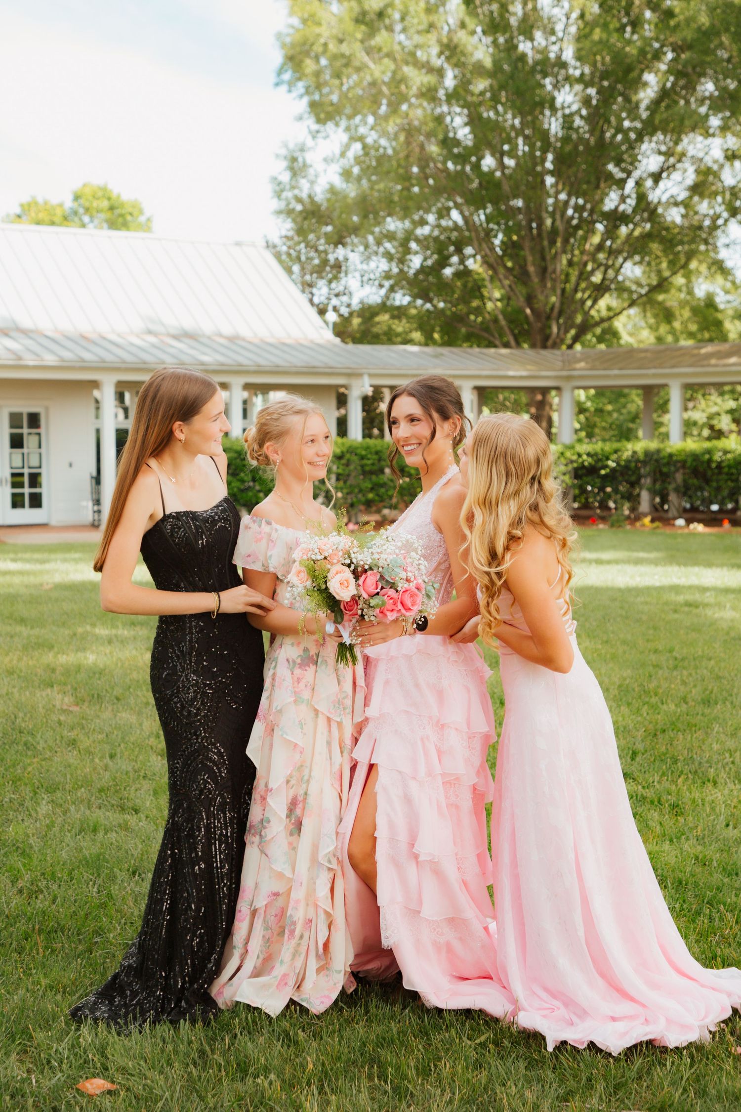 Four young women in formal gowns gathered together on green lawn, one holding pink bouquet near white venue.