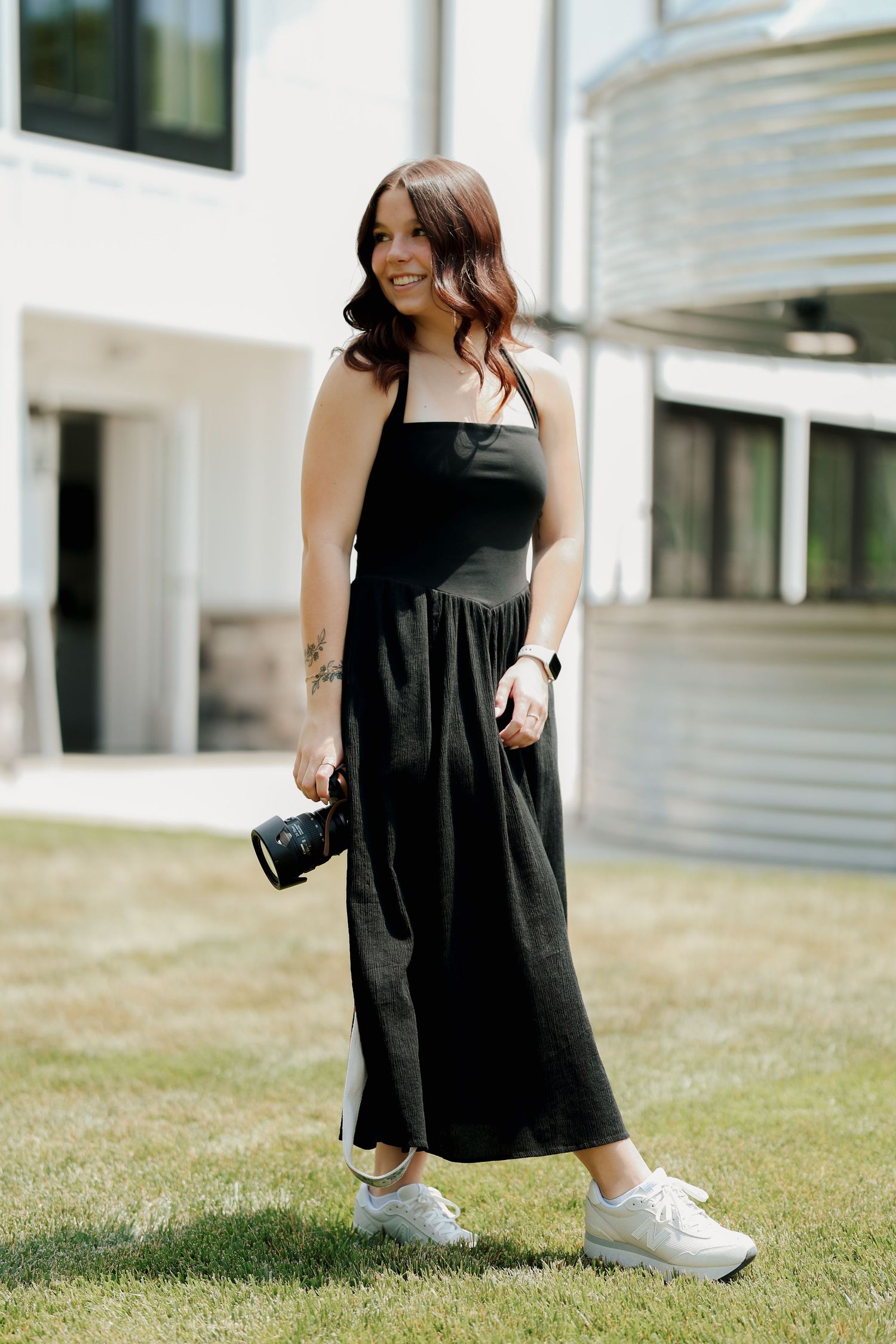 Brunette female photographer in black maxi dress walks on grass holding camera outdoors.
