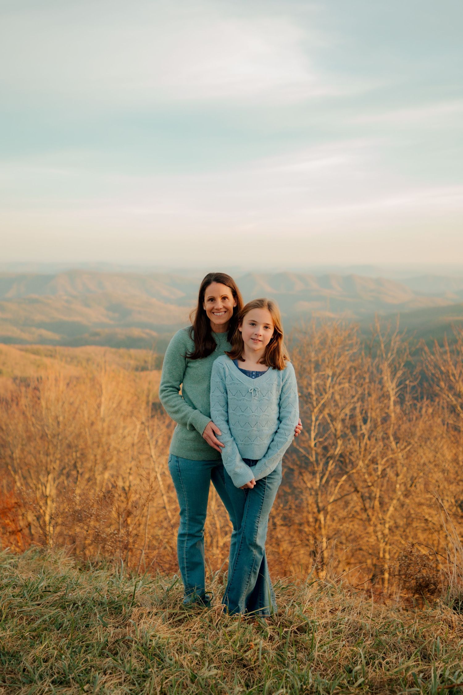 Mother and daughter stand together smiling with golden autumn mountain landscape in background.