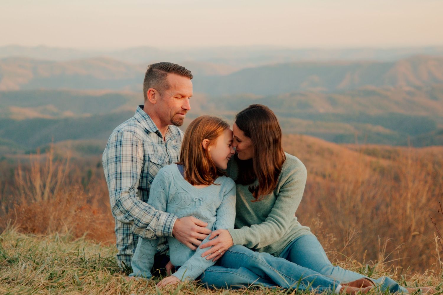A family of three shares a sweet nose-to-nose moment while seated on a hilltop with mountain scenery behind them.