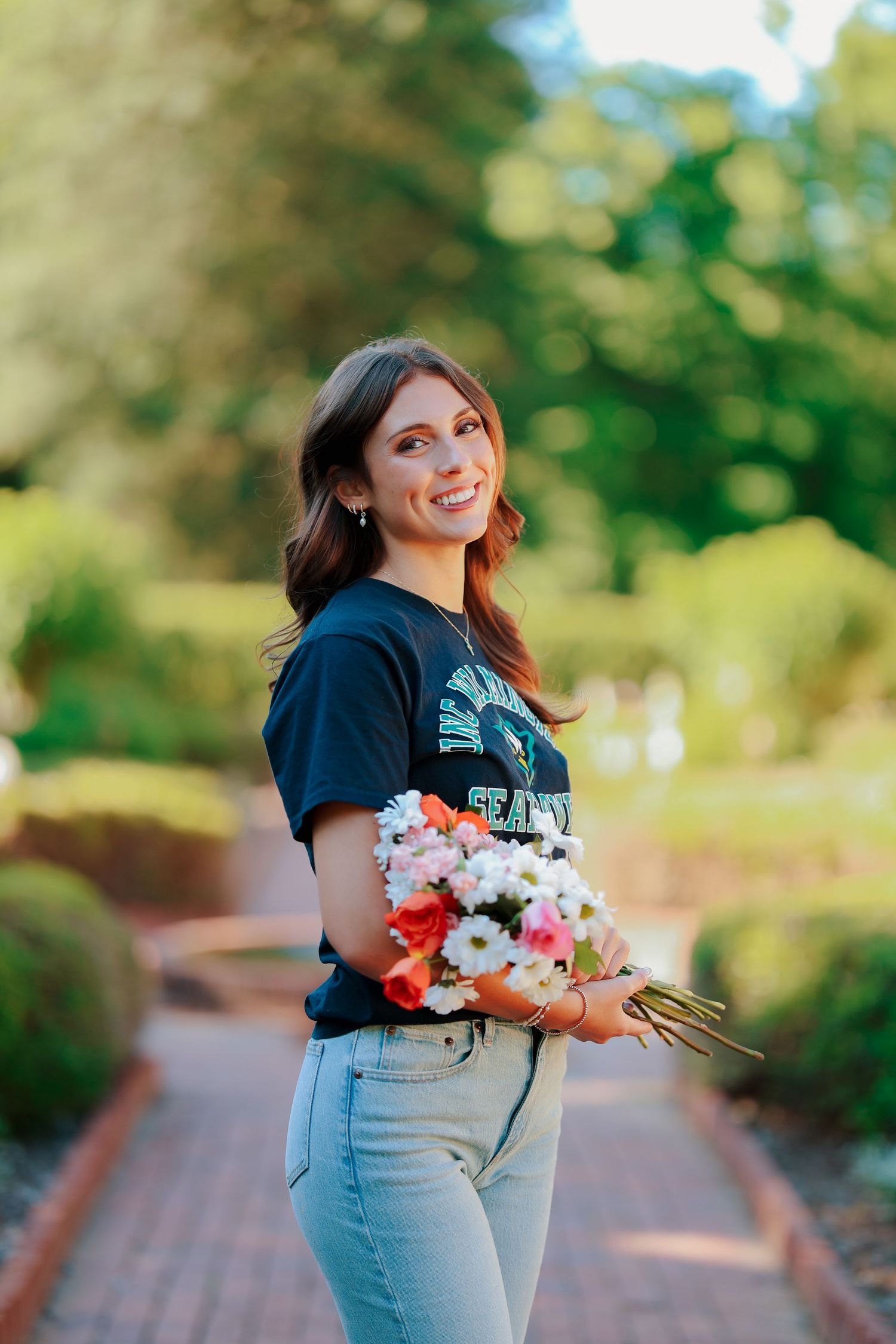 Smiling young woman holds a colorful bouquet of flowers on a brick garden path outdoors.
