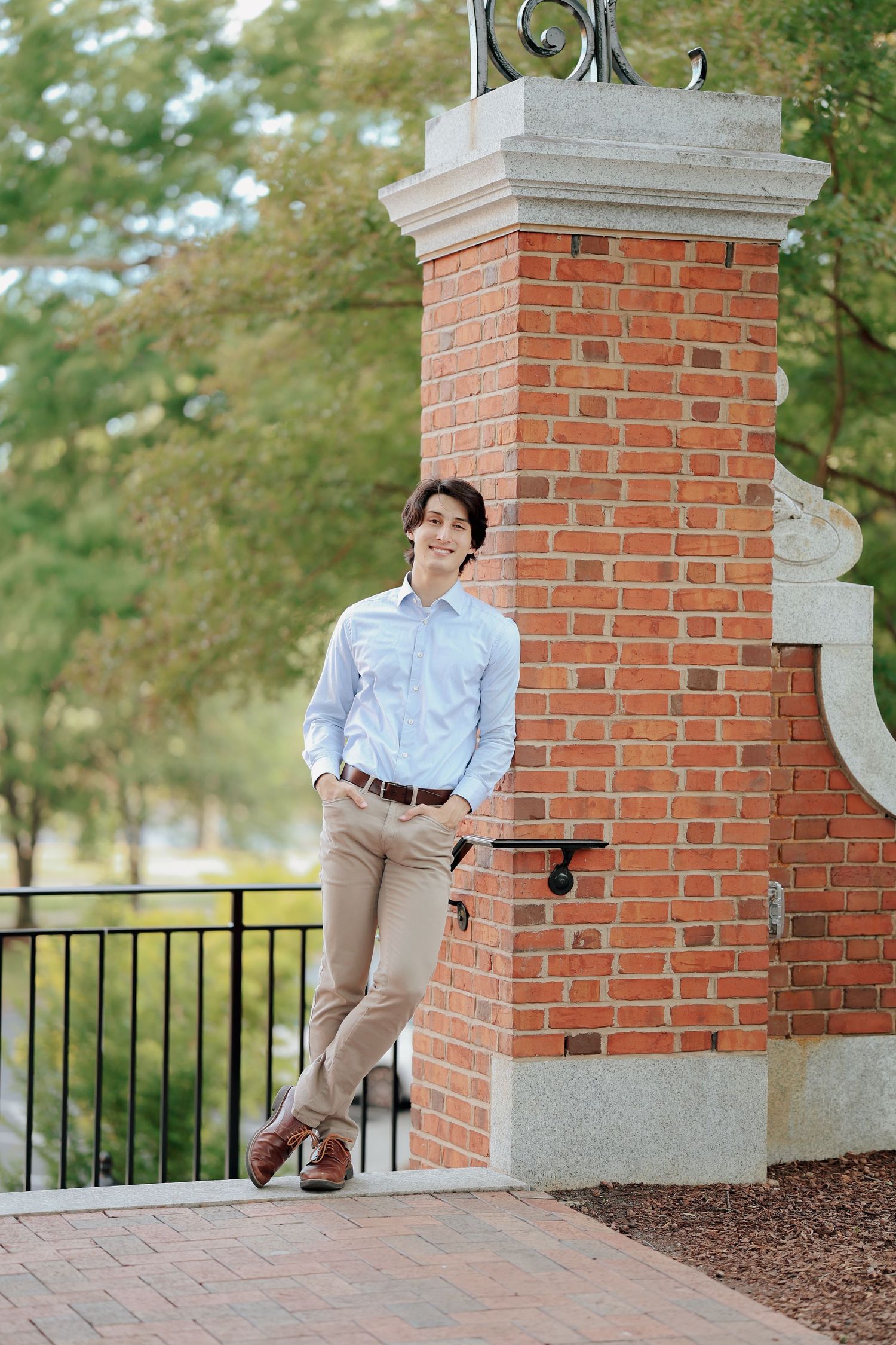 Smiling young man in blue shirt and khakis leans against a brick pillar at a campus gate.
