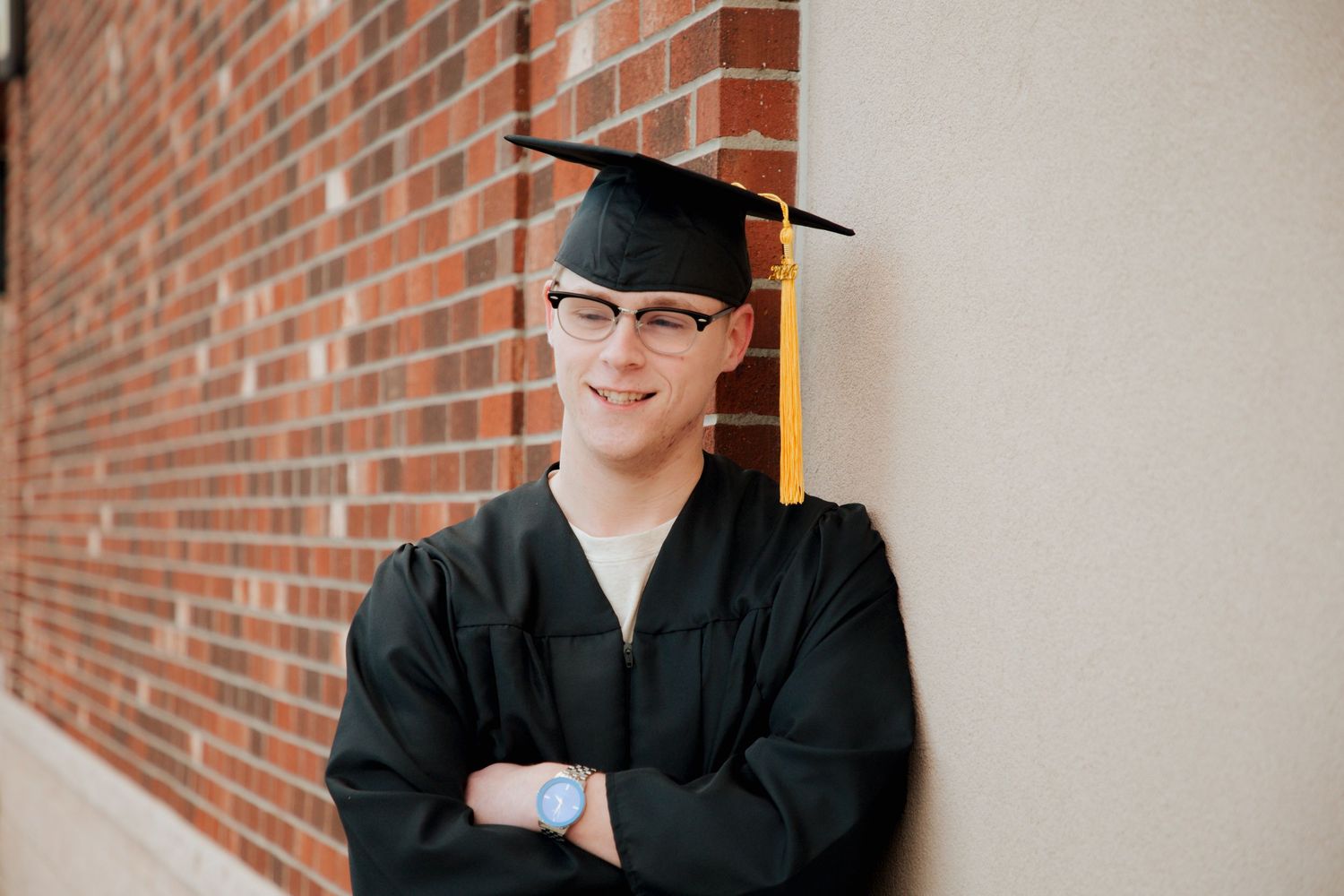 Smiling graduate in glasses wearing black cap and gown leaning against white wall.