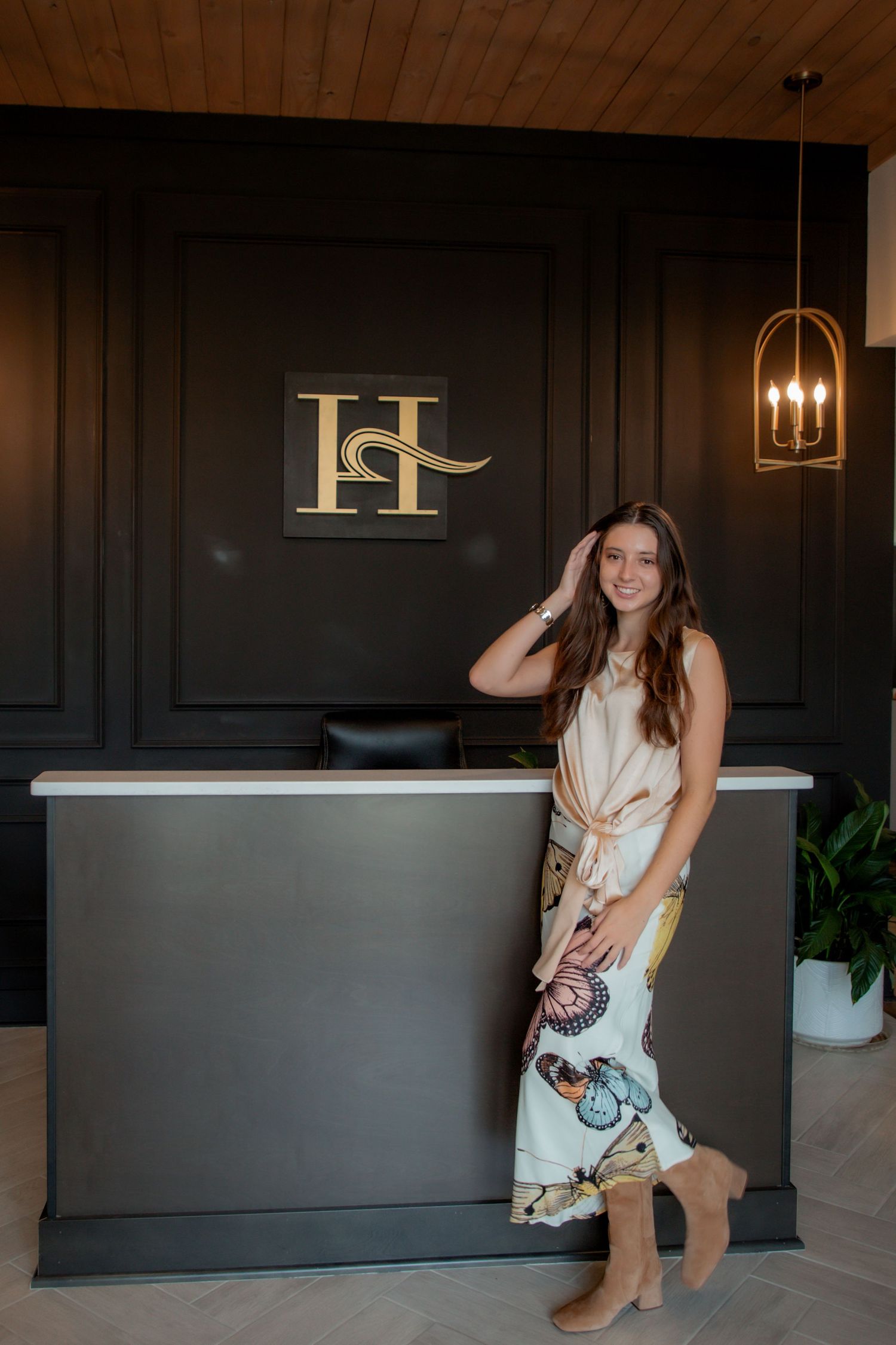 Young woman in butterfly skirt and beige top posing at elegant dark reception desk with gold H logo.