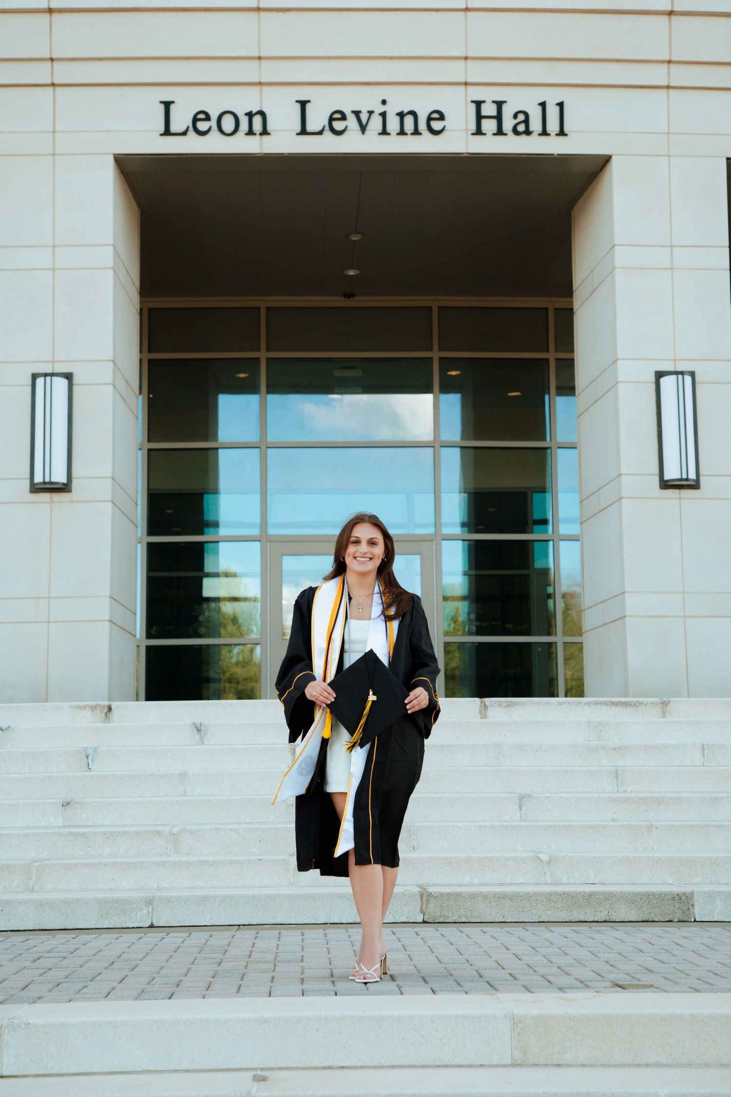 Smiling female graduate in black gown walks down steps of Leon Levine Hall.