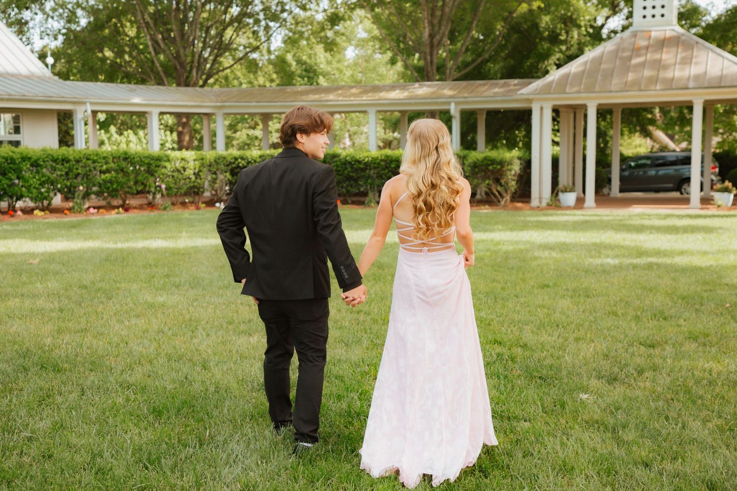 Teen prom couple walking hand in hand across green lawn toward white columned building.