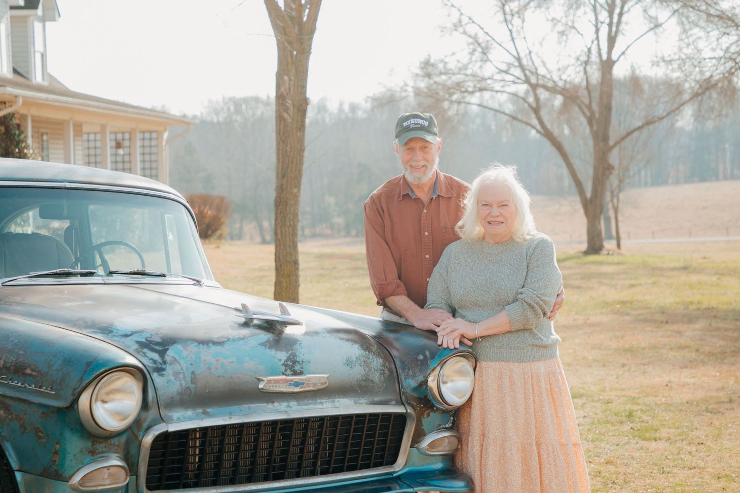 Elderly couple posing together beside a rustic vintage teal classic car on a sunny day near a farmhouse.
