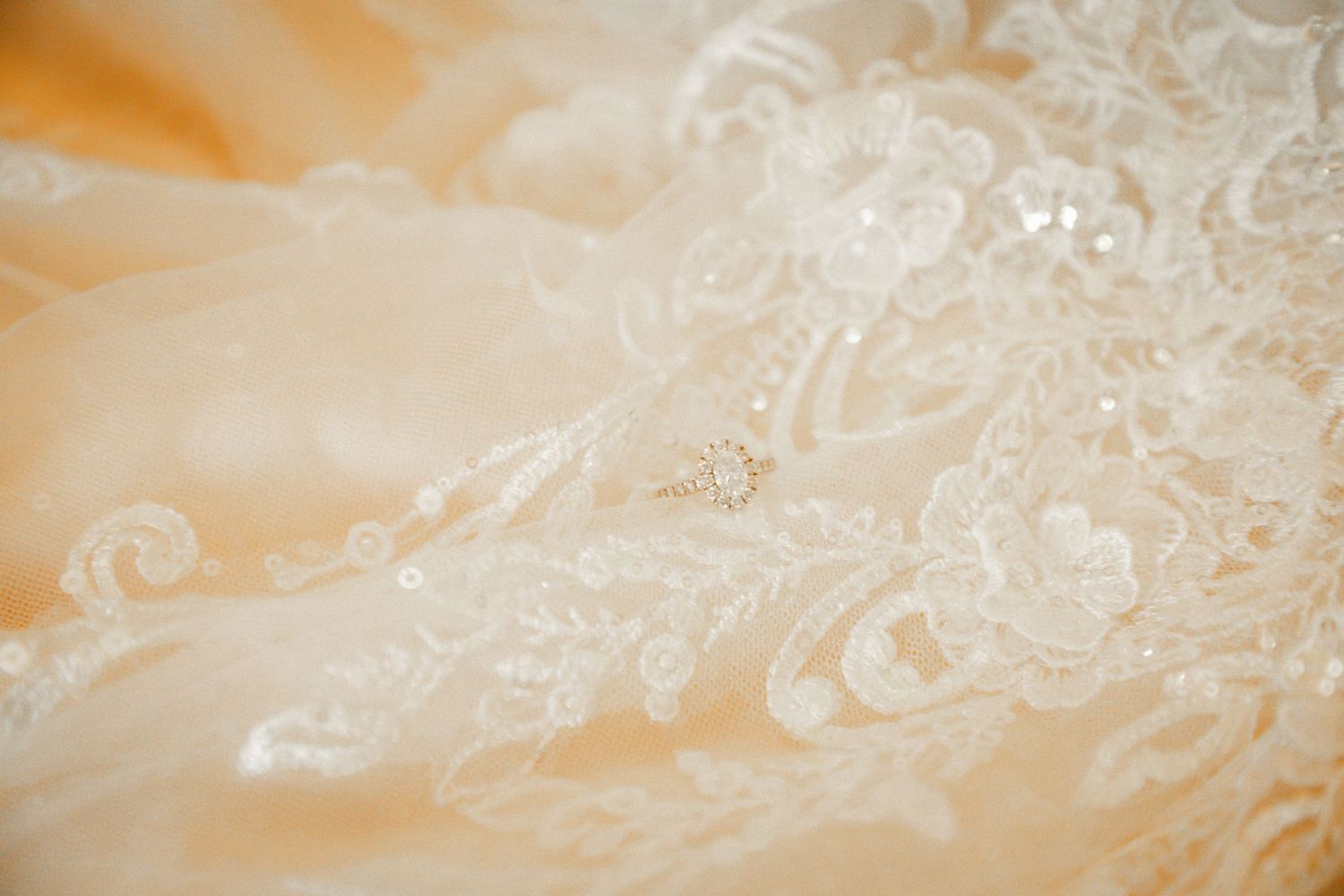 Diamond engagement ring resting on intricate ivory lace wedding dress fabric with floral embroidery.