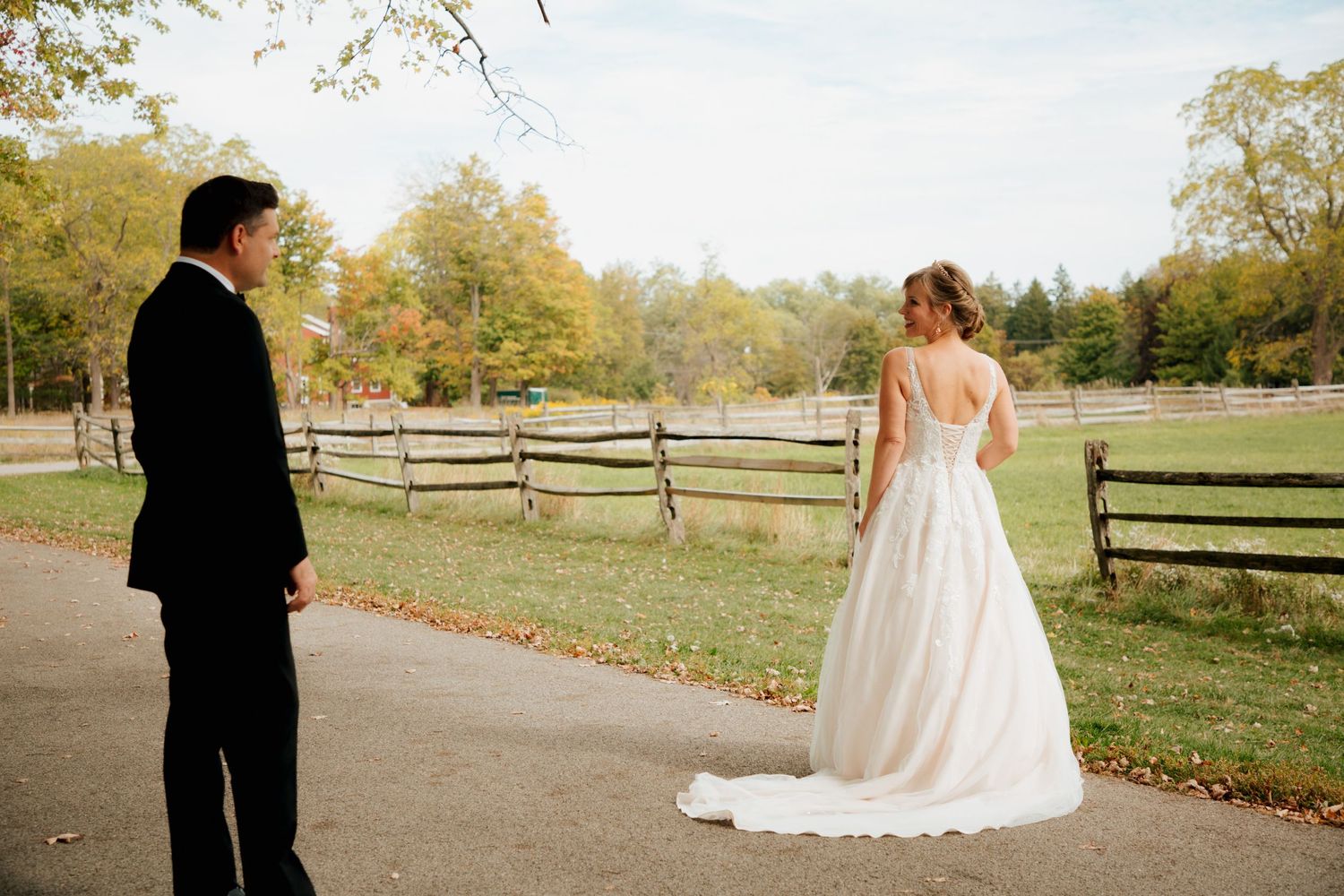 Groom watching bride in elegant white gown with train at rustic fall farm wedding.