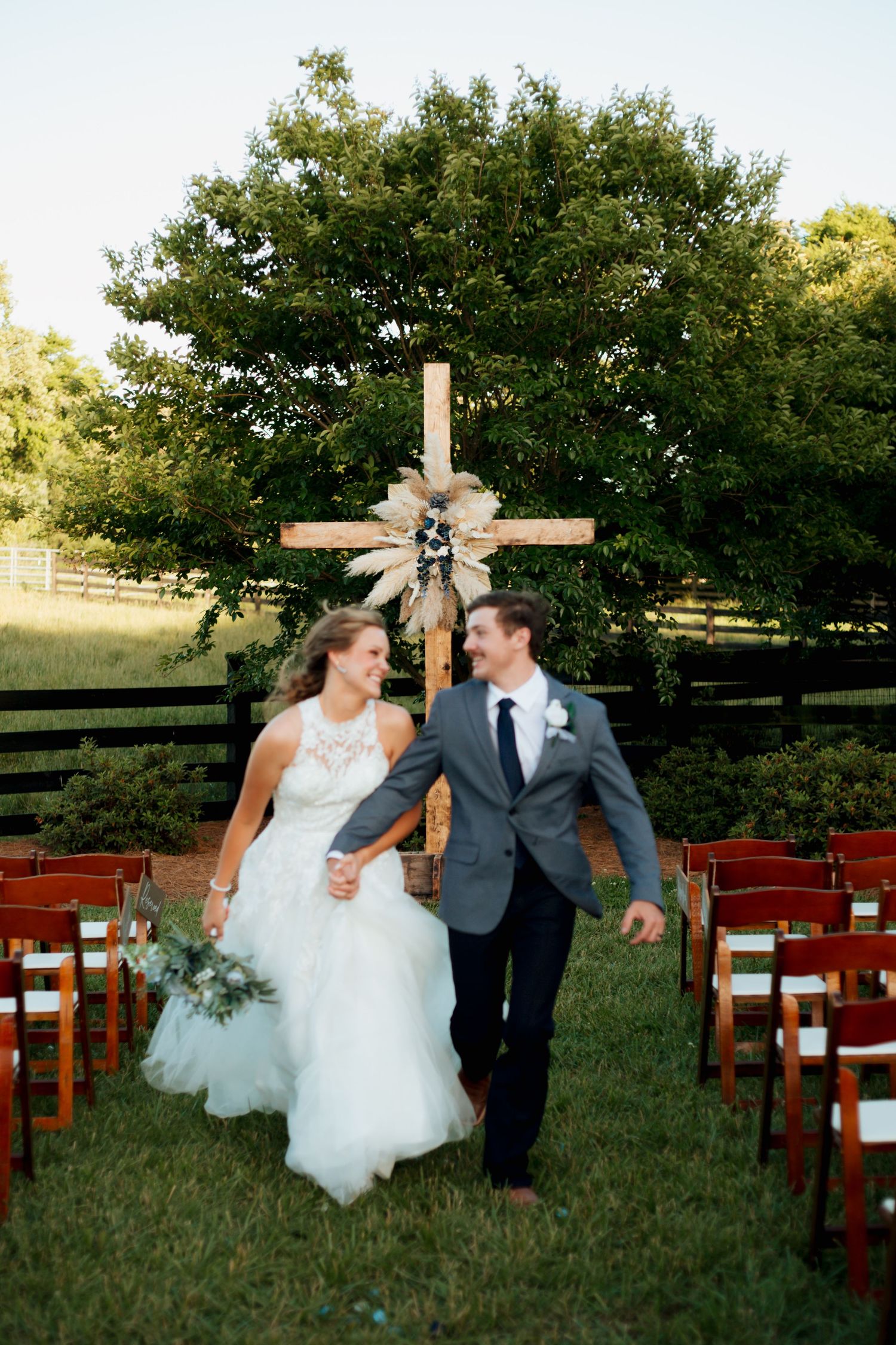 Newlyweds walk hand in hand down the aisle past wooden chairs at an outdoor rustic farm wedding ceremony.