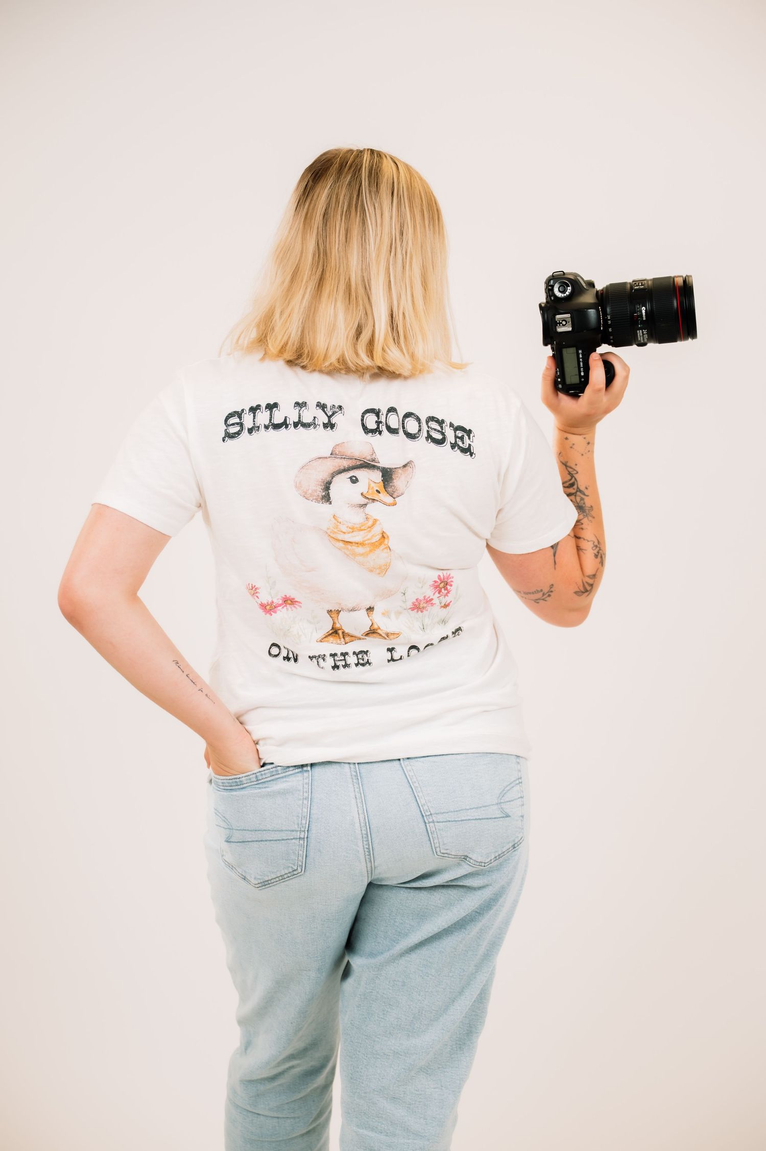 Back view of woman in Silly Goose On The Loose graphic tee holding DSLR camera in studio.